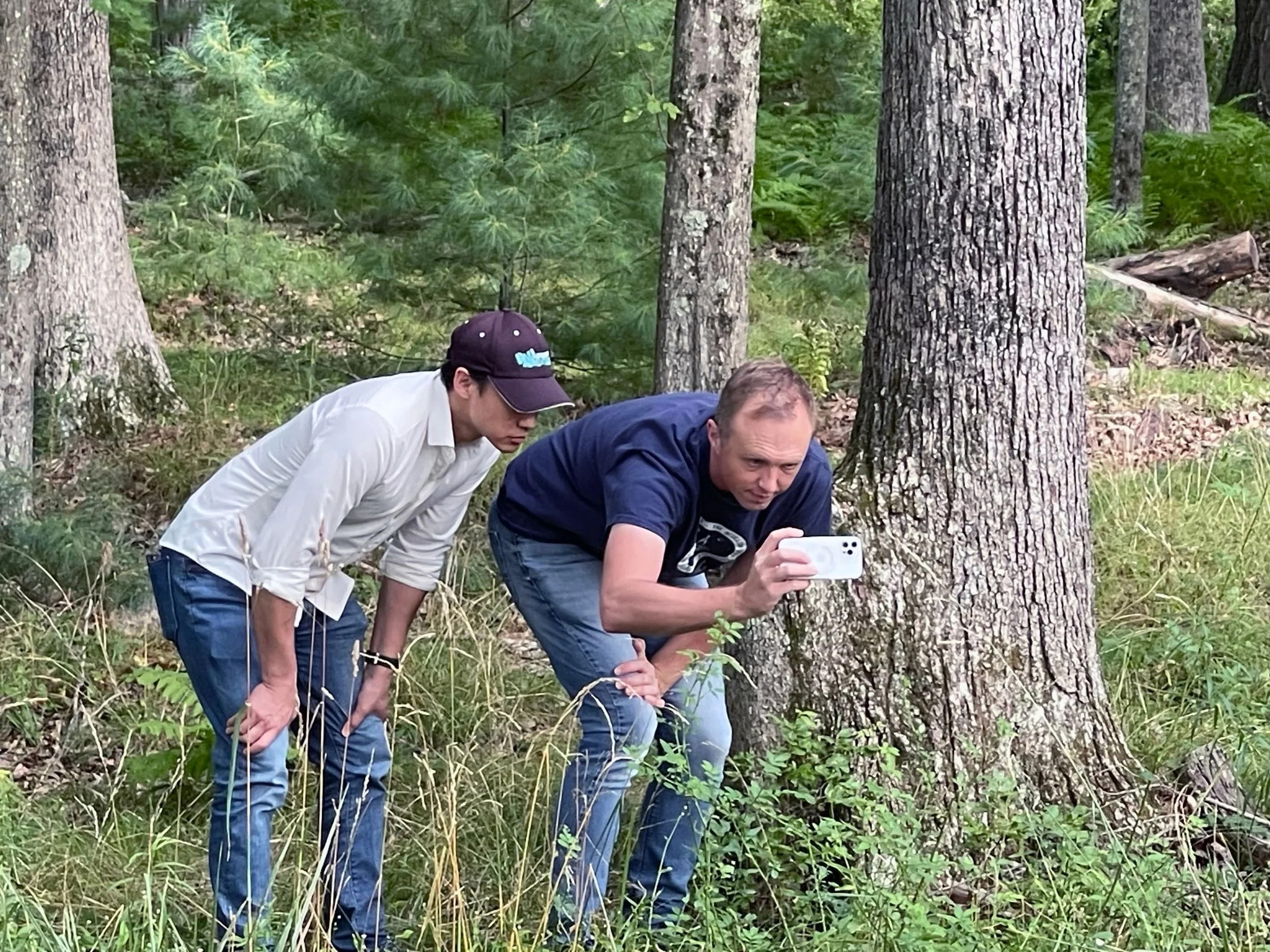 Director Joe Lam and cinematographer Jeremy Royce crouching together in a forest, reviewing a shot on a phone during a location tech scout for The Fetus