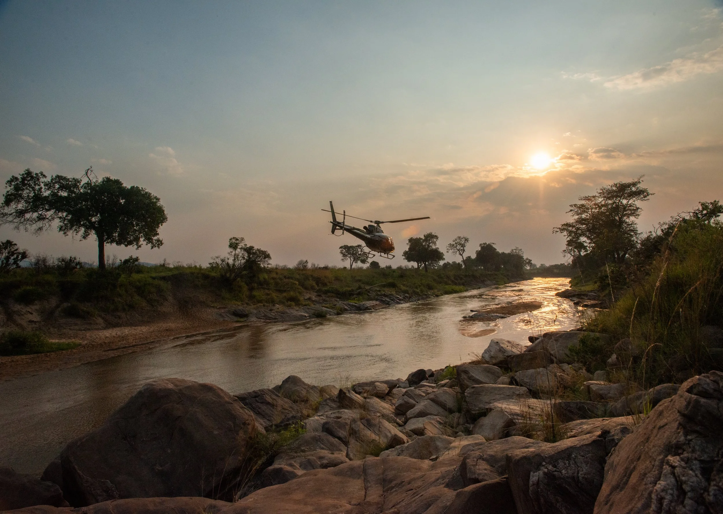 A helicopter flying over a river at sunset surrounded by trees and rocks.