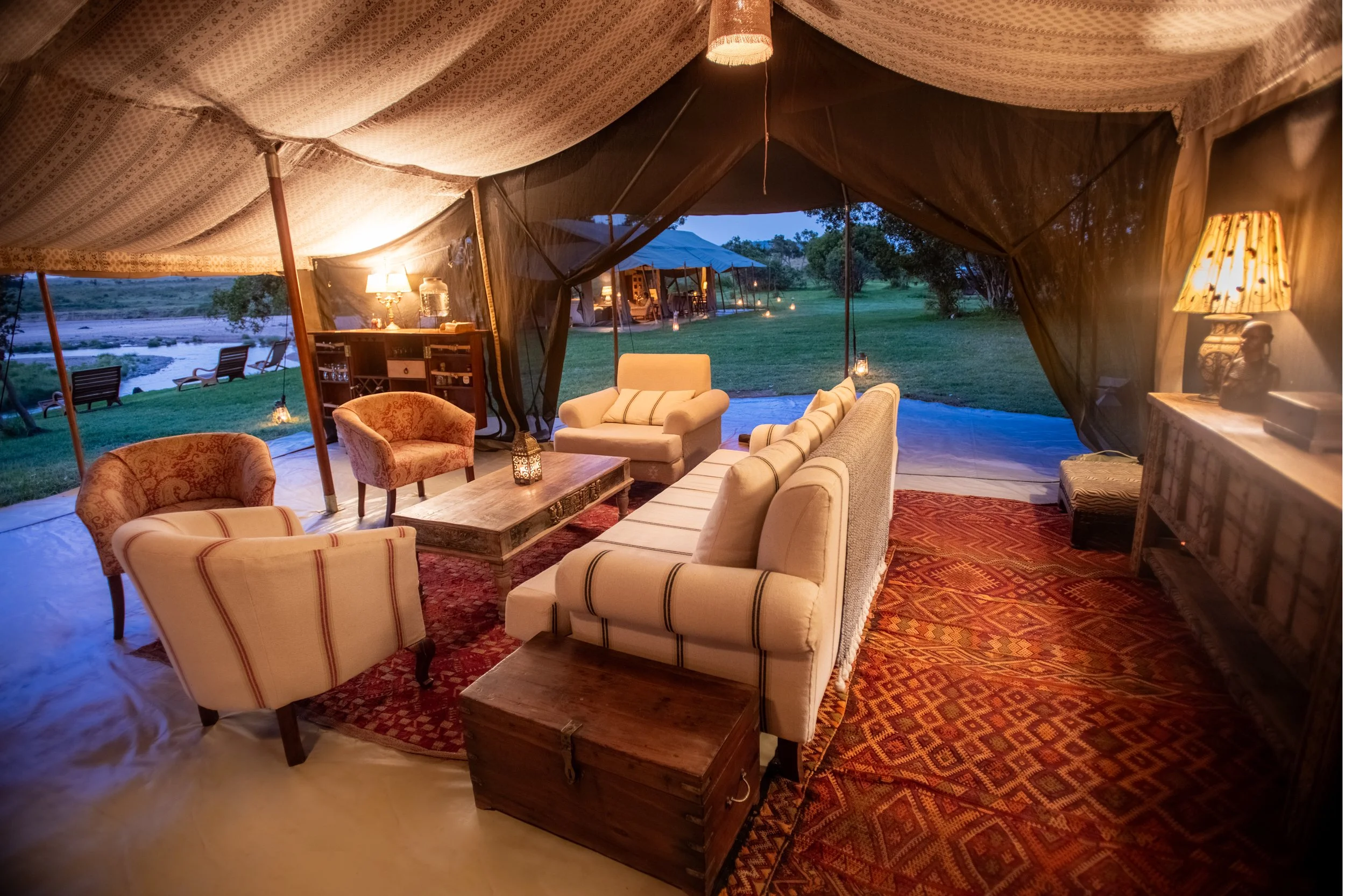 Interior of a luxurious tent with cozy seating, including a sofa, armchairs, and a coffee table, overlooking a scenic outdoor landscape with trees and a river at dusk.