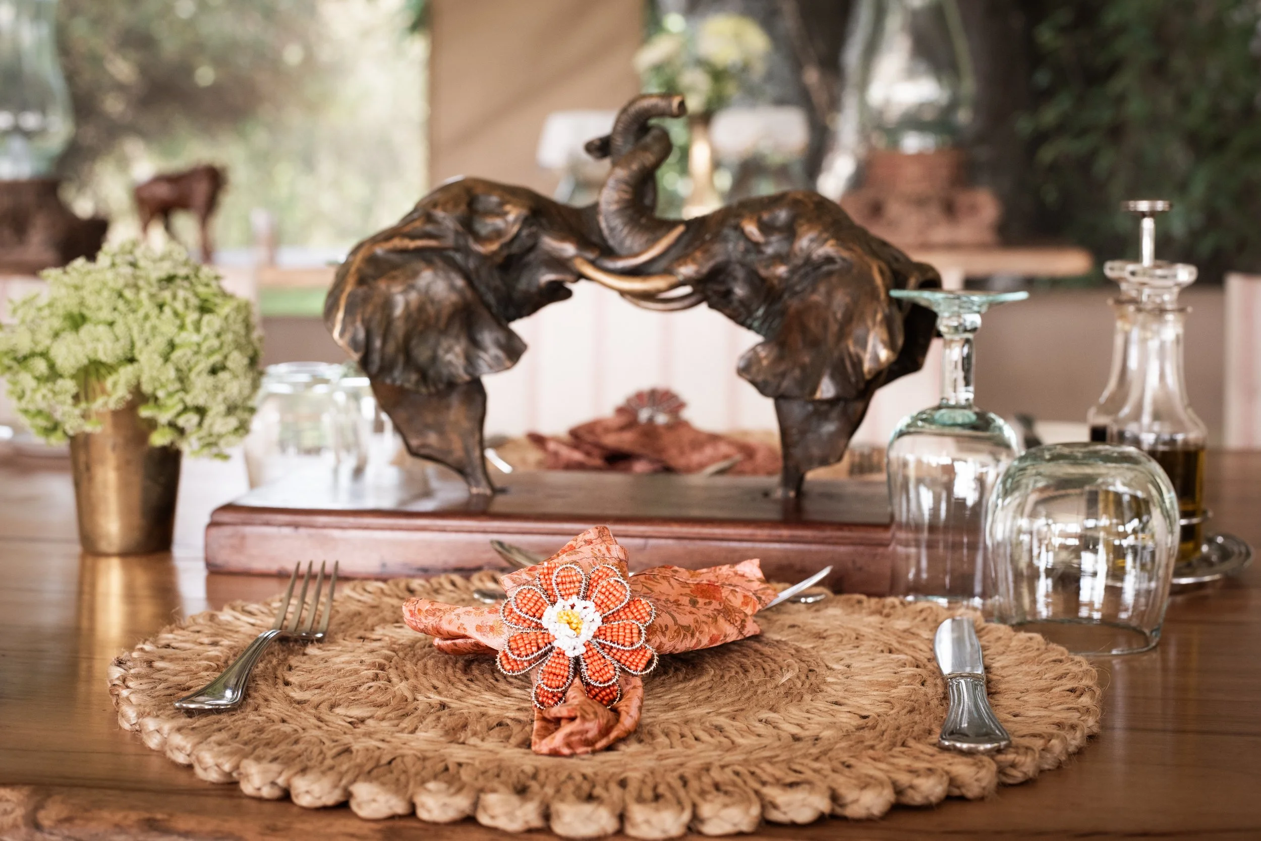 A rustic dining table set with a woven placemat, silverware, a cloth napkin with a beaded flower decoration, a small potted green plant, and decorative glass bottles. In the background, there is a sculpture of two elephants with their trunks intertwi