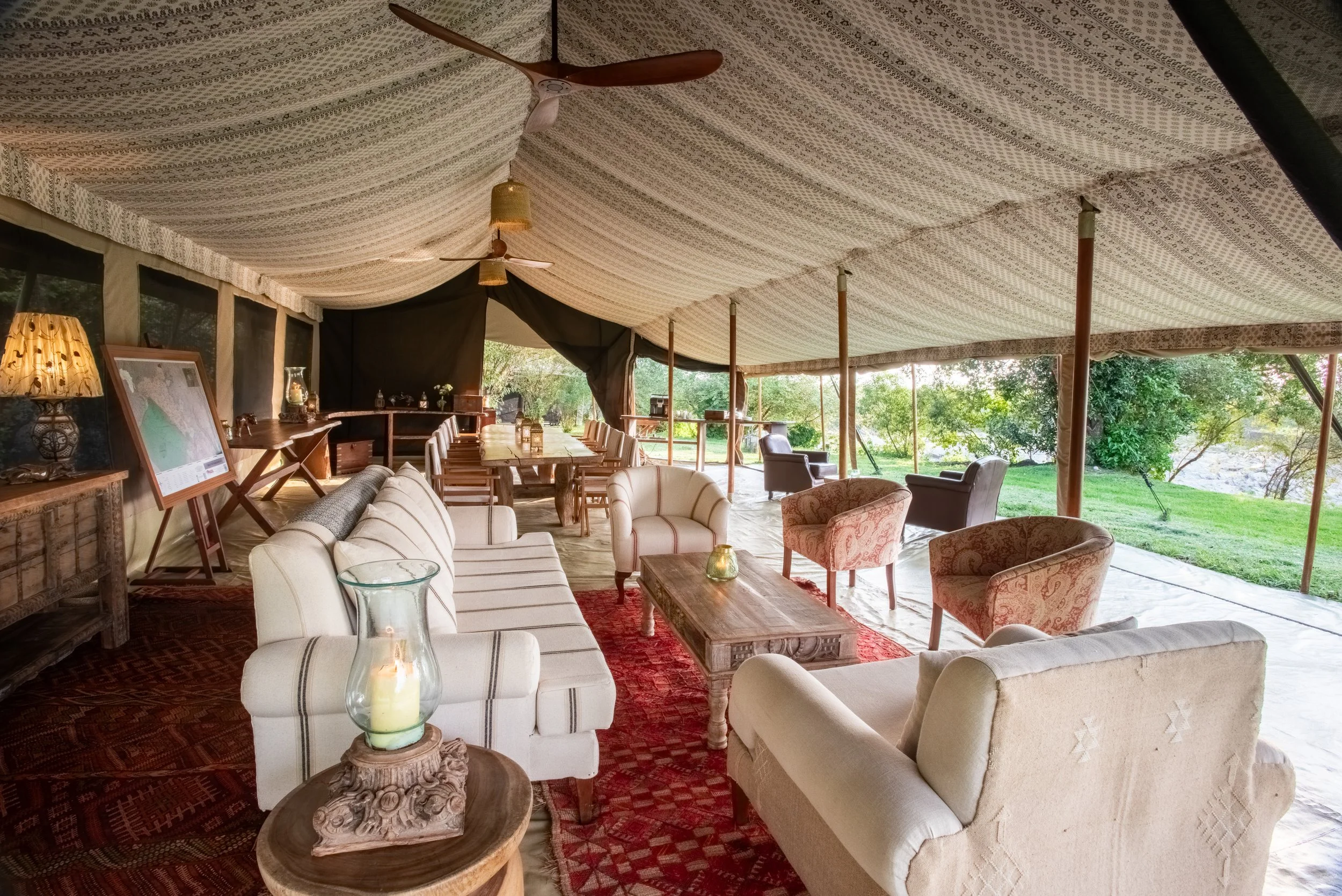 Interior of a spacious tent set up for social gathering, featuring sofas, armchairs, a large dining table, and decorative lamps, with an outdoor view of greenery.