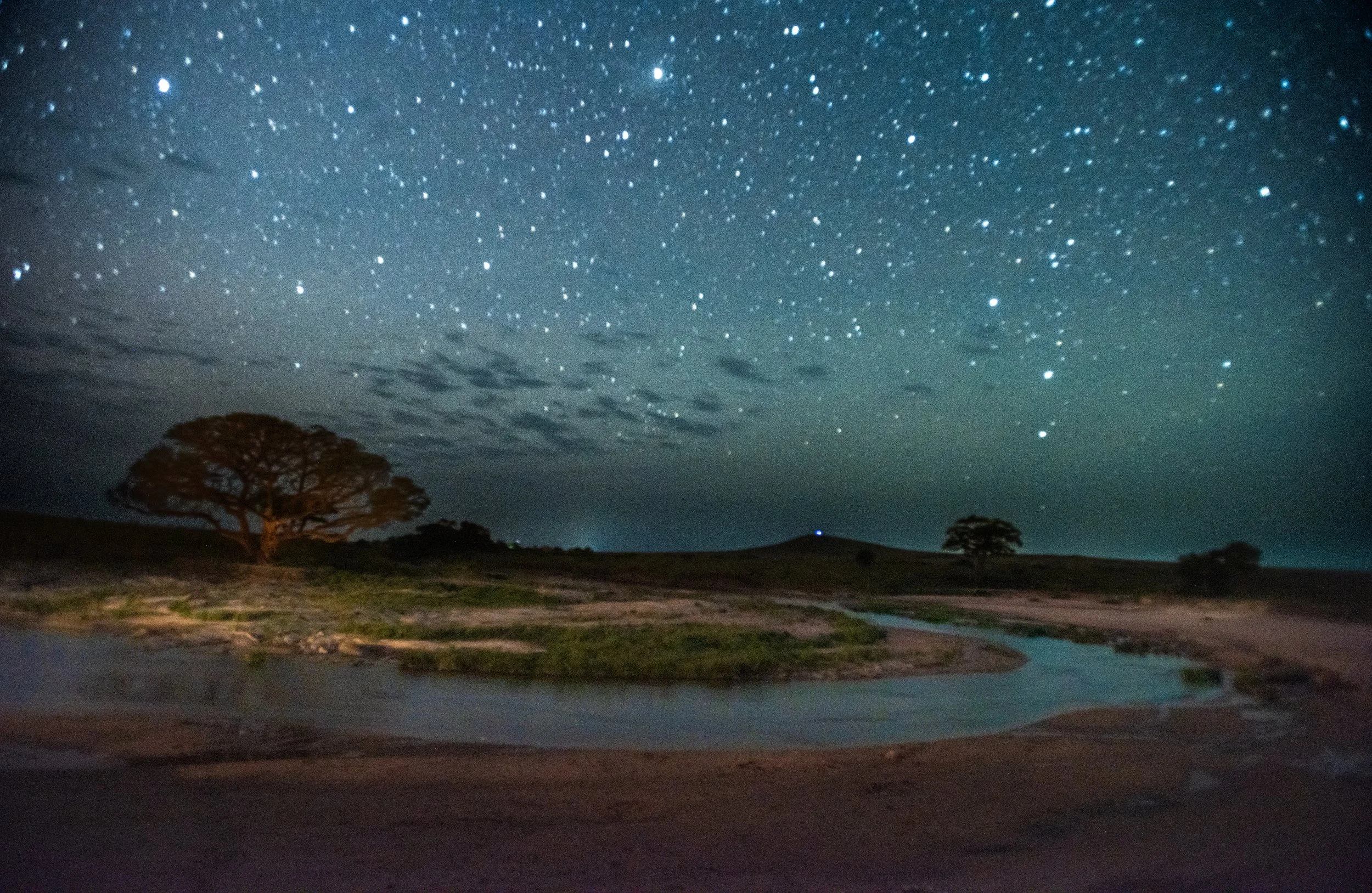 Nighttime landscape with stars, a calm river, and trees on a hilly terrain.