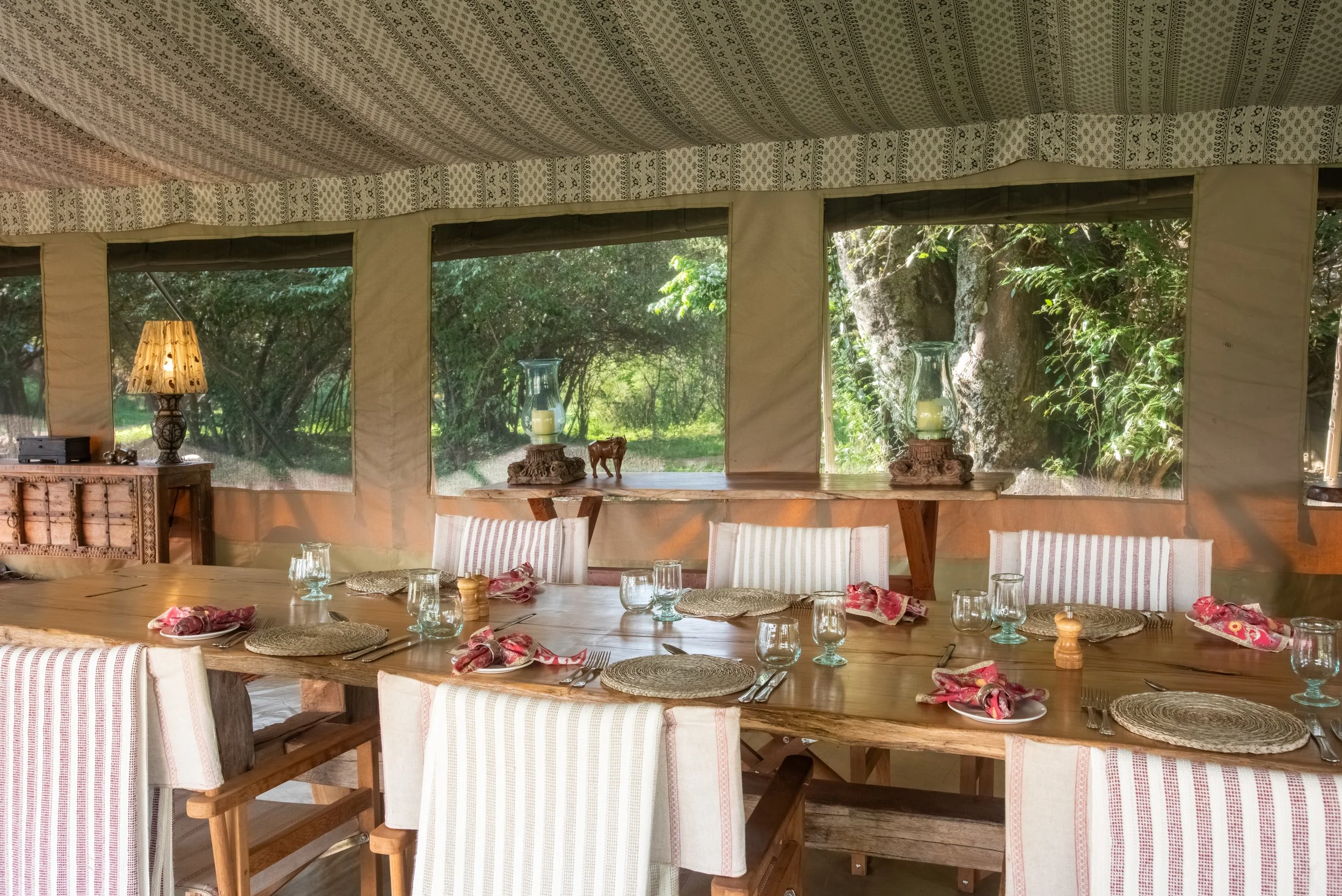 Dining room with large wooden table, striped upholstered chairs, and a view of lush green trees outside through windows. The table is set with woven placemats, glasses, and cloth napkins.