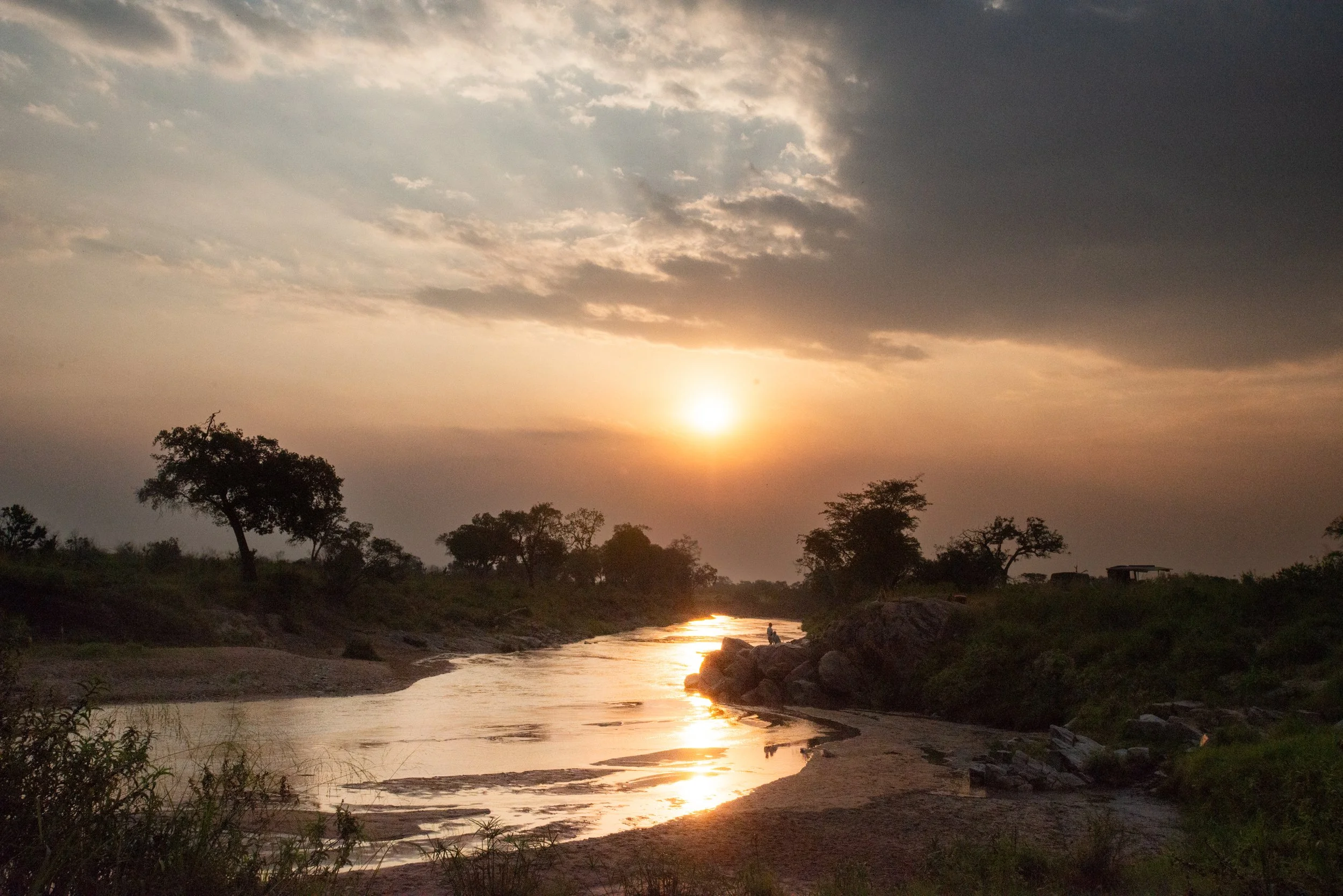 Sunset over a river with silhouettes of trees and rocks along the bank.
