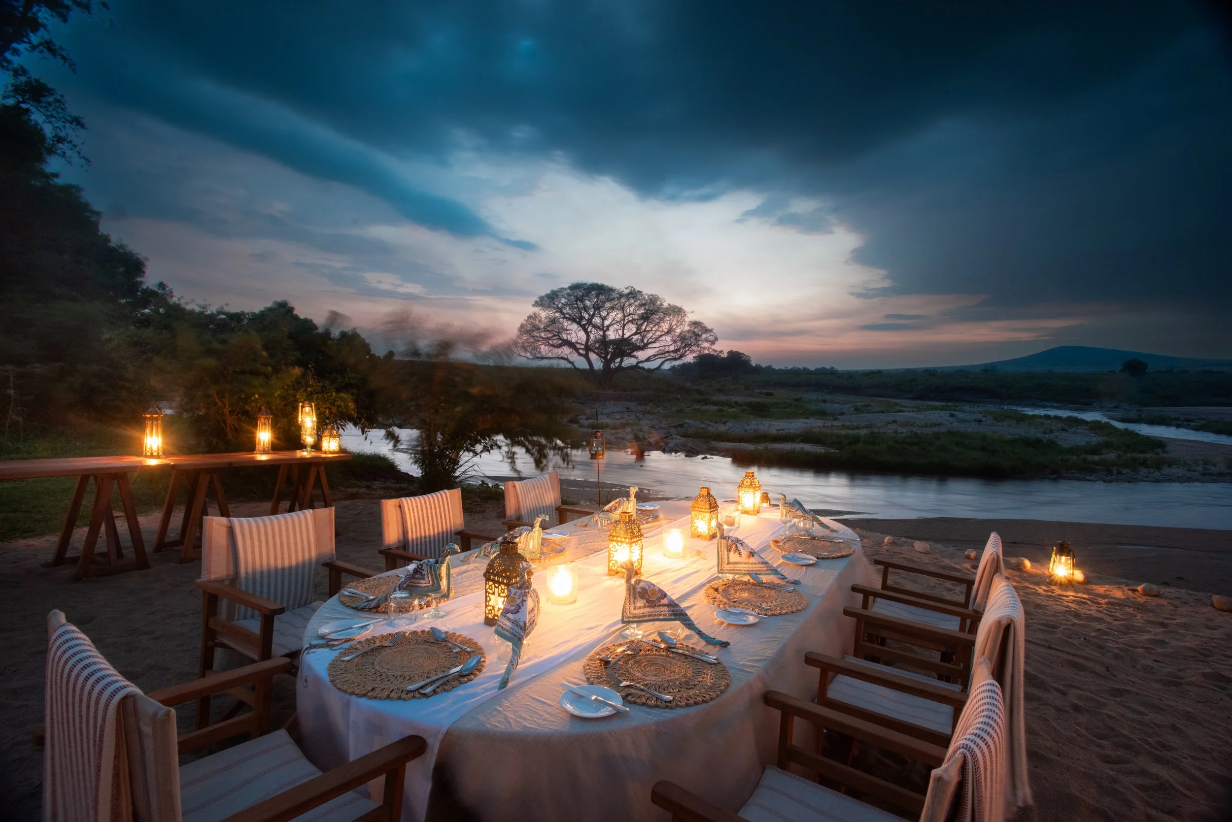 Outdoor dining setup at sunset or dusk by a river, with a long table decorated with lanterns, plates, and napkins, surrounded by wooden chairs, and a scenic landscape with trees and hills in the background.