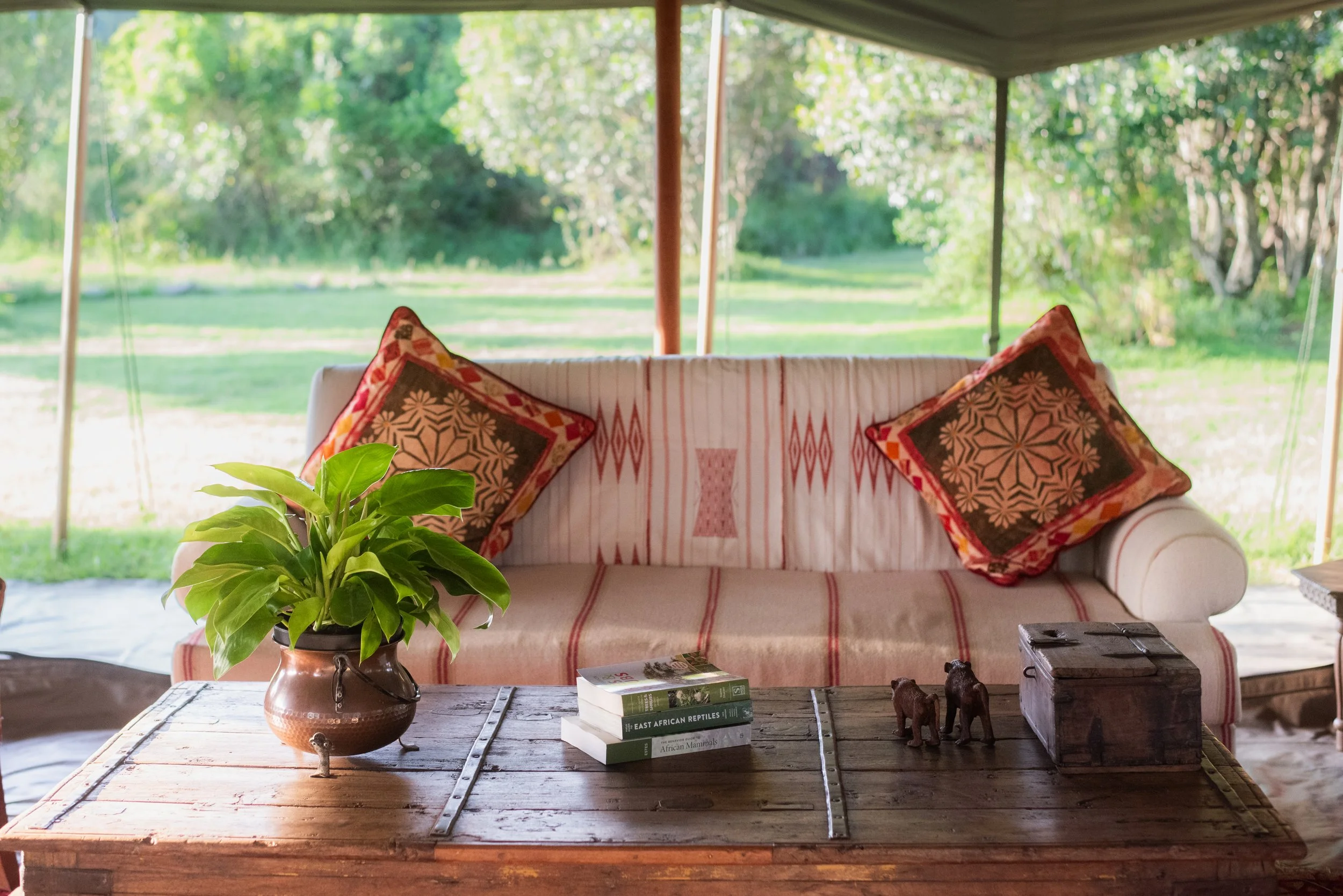 Living room with a white sofa, two decorative pillows with red and black patterns, a wooden coffee table with a potted green plant, books, and small animal figurines, with a view of green trees and grass outside through large open windows.