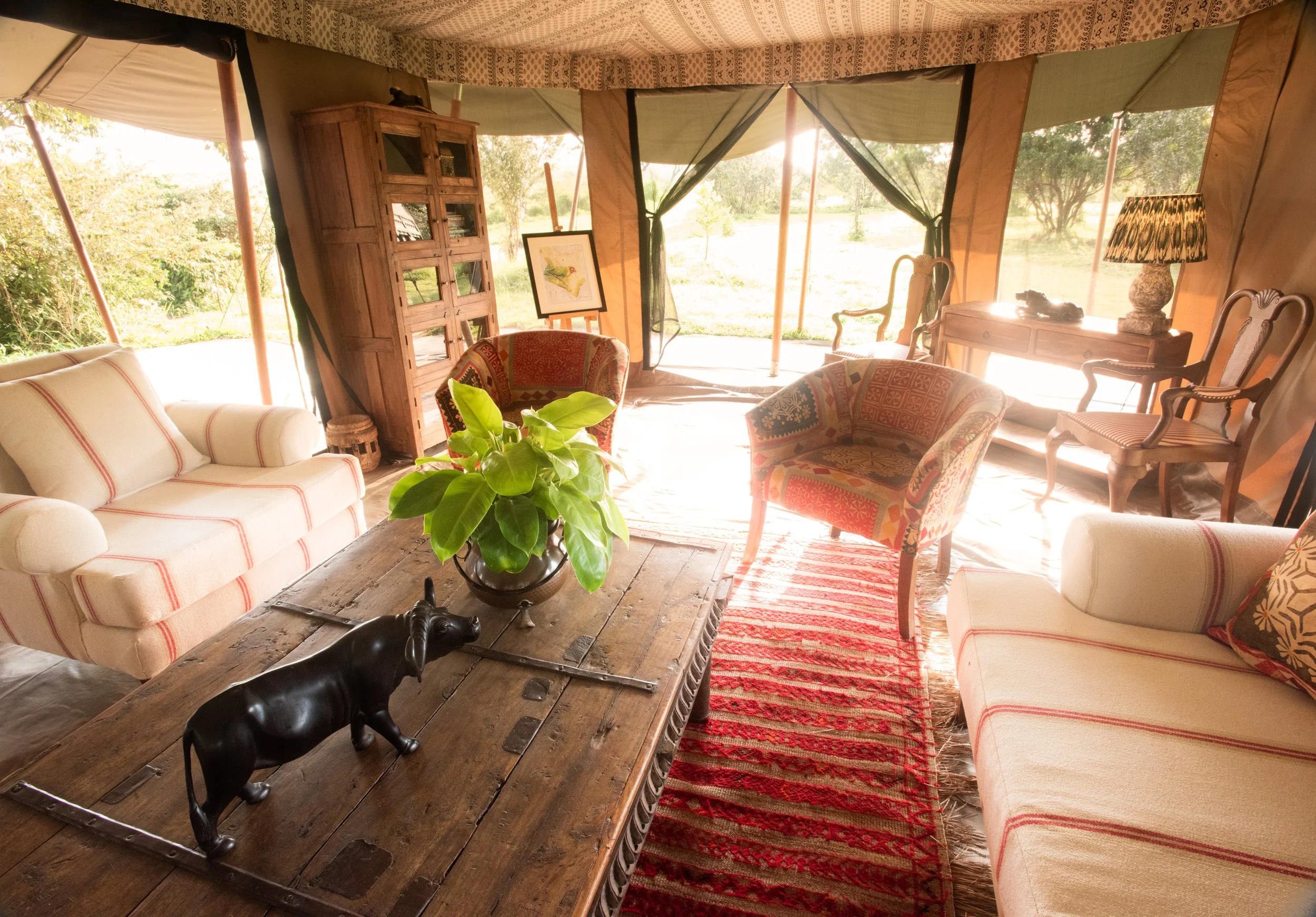 Cozy living room with vintage furniture, a wooden table with a black bull sculpture and a green potted plant, colorful patterned armchairs, a red striped area rug, and large windows with black curtains showing greenery outside.