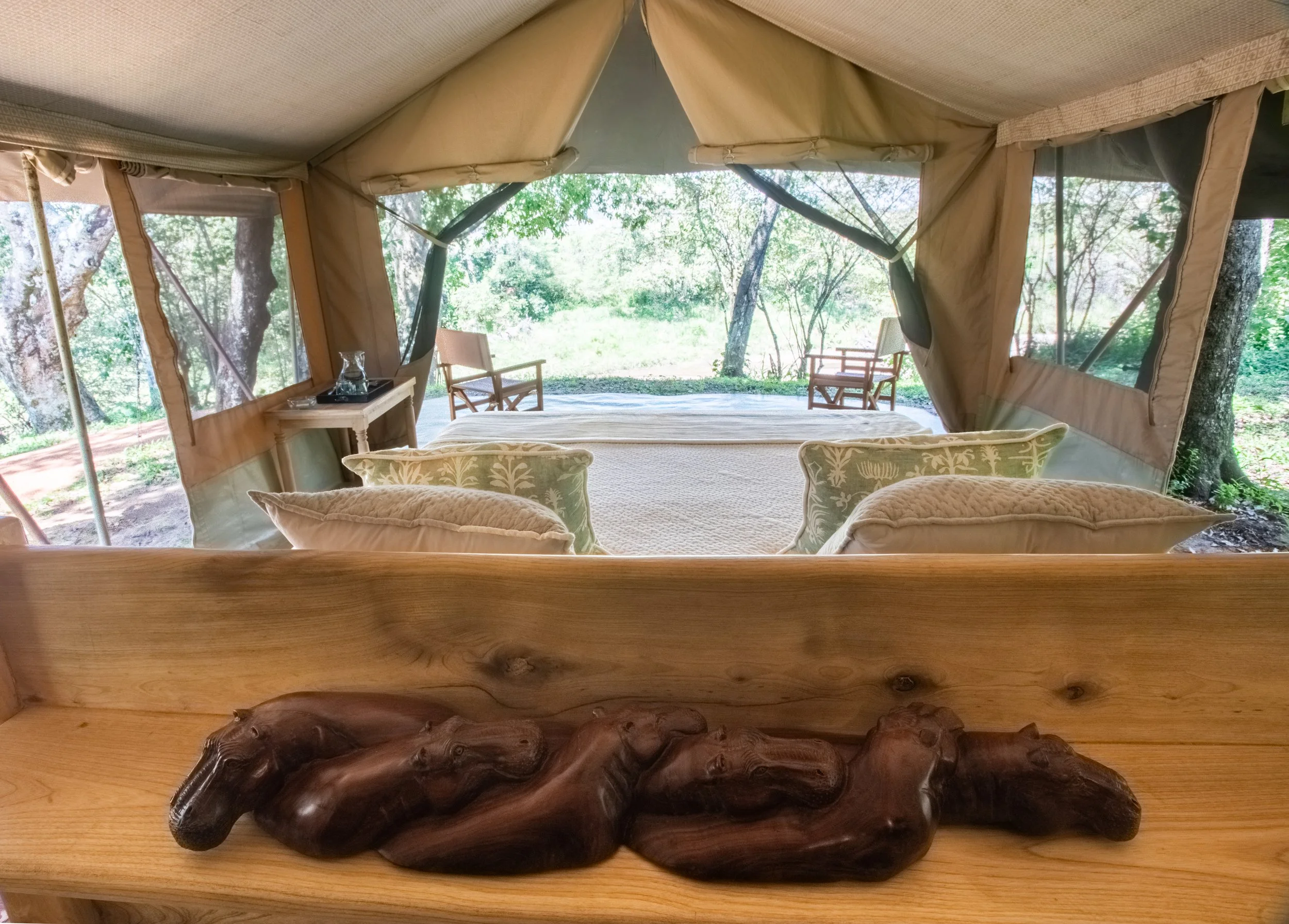 Interior of a luxury outdoor tent with a bed, decorative pillows, and a view of trees and an outdoor sitting area with chairs.