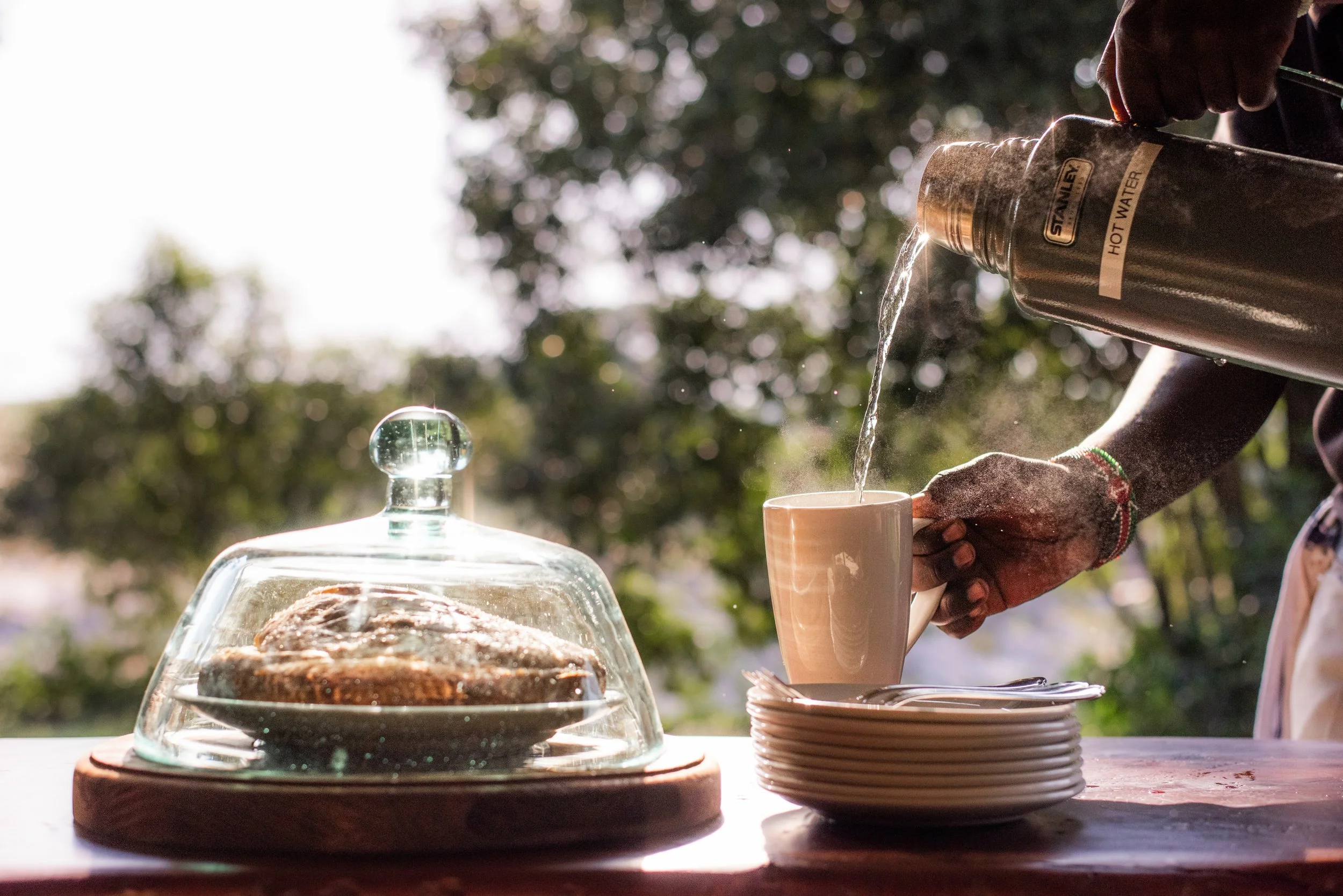 Person pouring hot water from a thermos into a white coffee cup on a stack of plates outdoors, with a glass cake dome covering a pastry on the left, sunlight filtering through trees in the background.