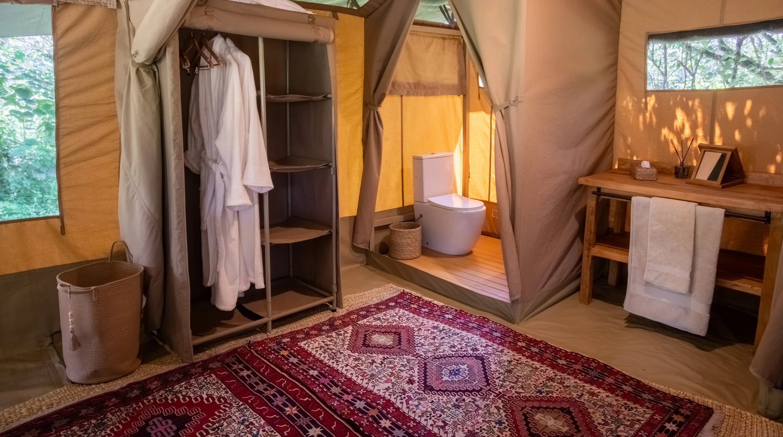 Interior of a rustic-style tent with a clothing rack holding white robes, a small bathroom with a toilet, a side table with a framed mirror, a tissue box, a small plant, and a towel, and a patterned area rug on the wooden floor.