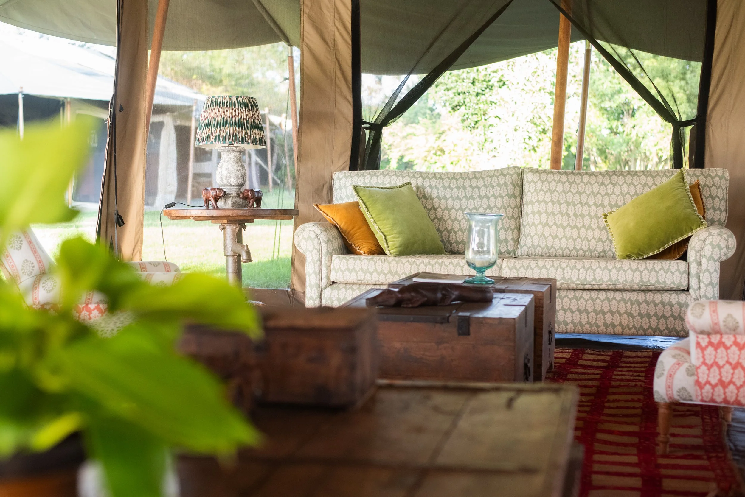 A cozy living room inside a large tent with a patterned sofa, colorful cushions, a wooden coffee table with decorative items, and a side table with a lamp showing a nature view outside through the tent's open sides.