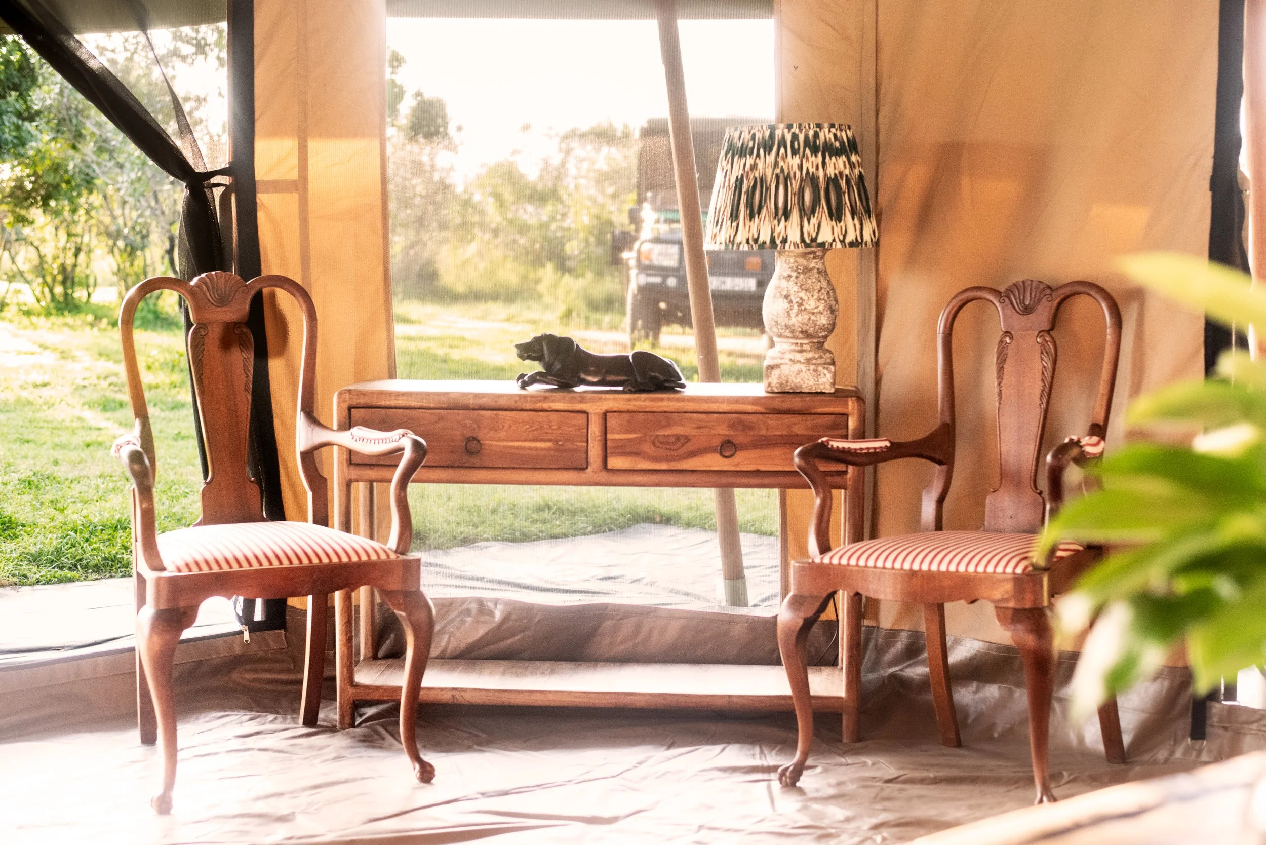 Interior of a tent with two wooden armchairs with striped red and white cushions, a wooden table with a black dachshund figurine, a lamp with a patterned shade, and a view of a grassy yard and vehicle outside.