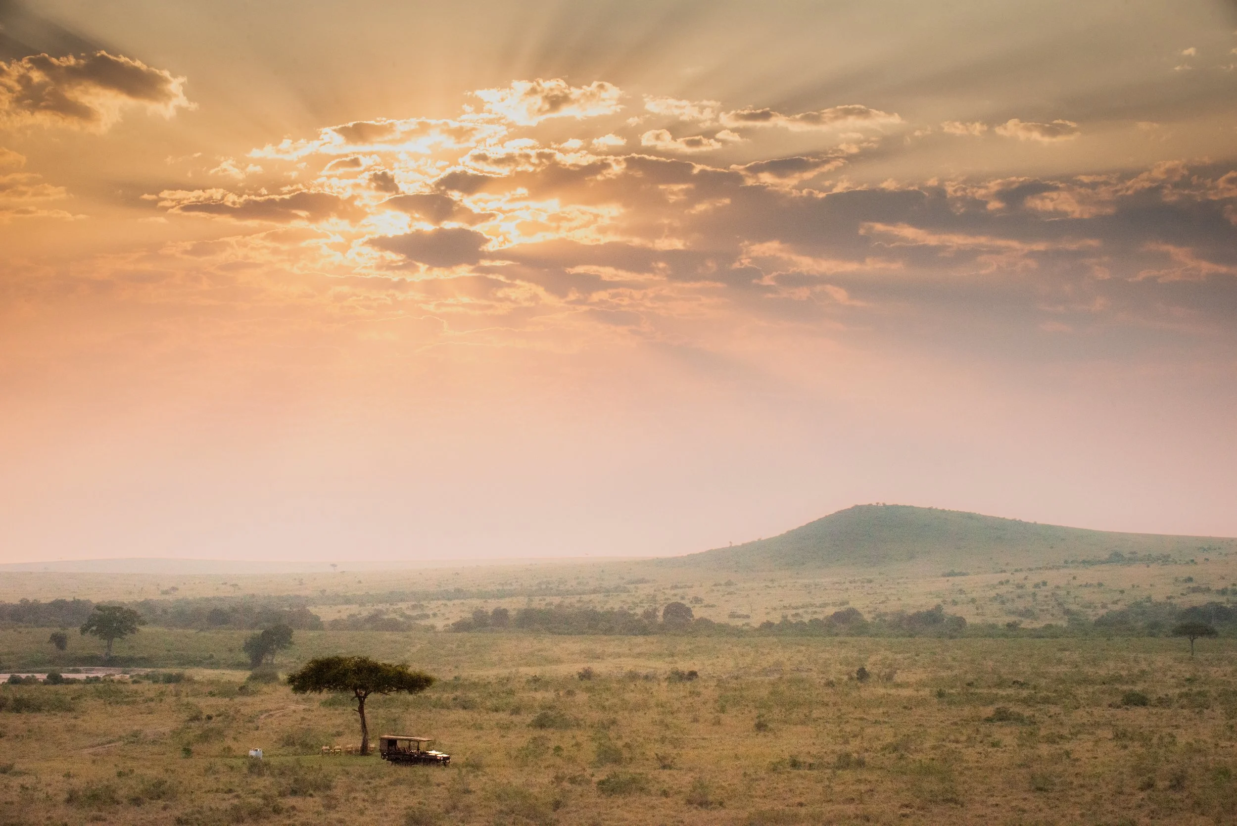 A vast savannah landscape at sunset with a few scattered trees, a small vehicle, and a hill in the distance under a sky with clouds and sunlight.