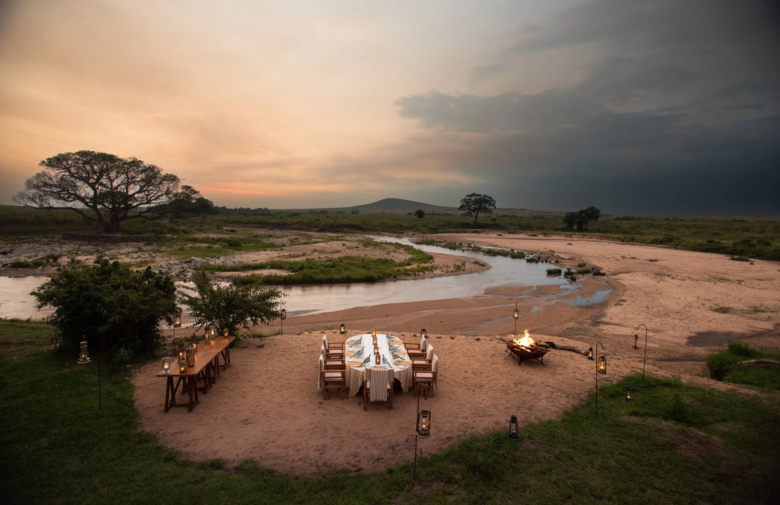 Dinner setup on a sandy riverbank during sunset with a long table surrounded by chairs, lanterns, and a campfire, overlooking a winding river and open landscape with trees and hills in the background.