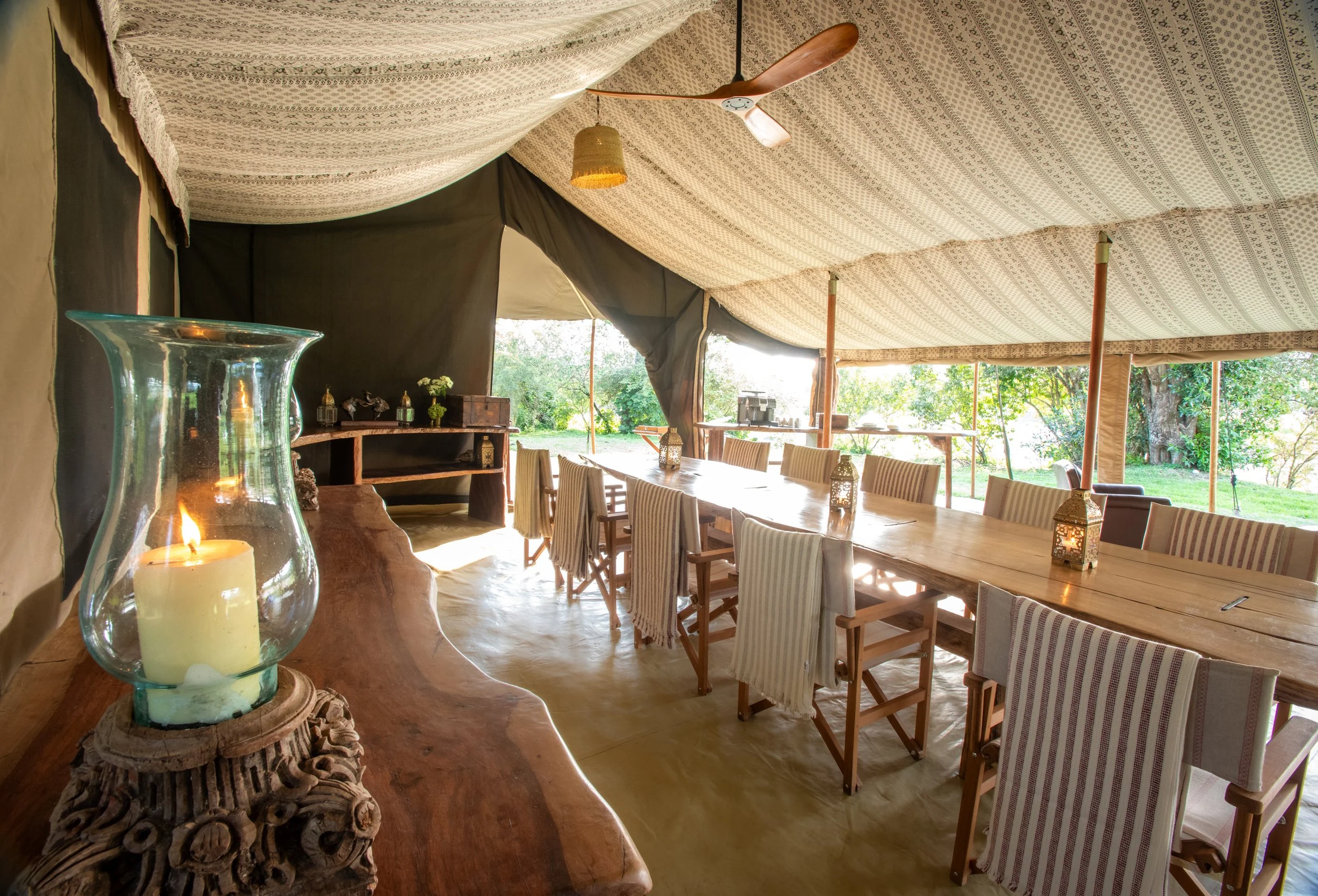 Tent interior with a long wooden dining table surrounded by striped chairs, decorated with lanterns, with a view of trees and greenery outside.