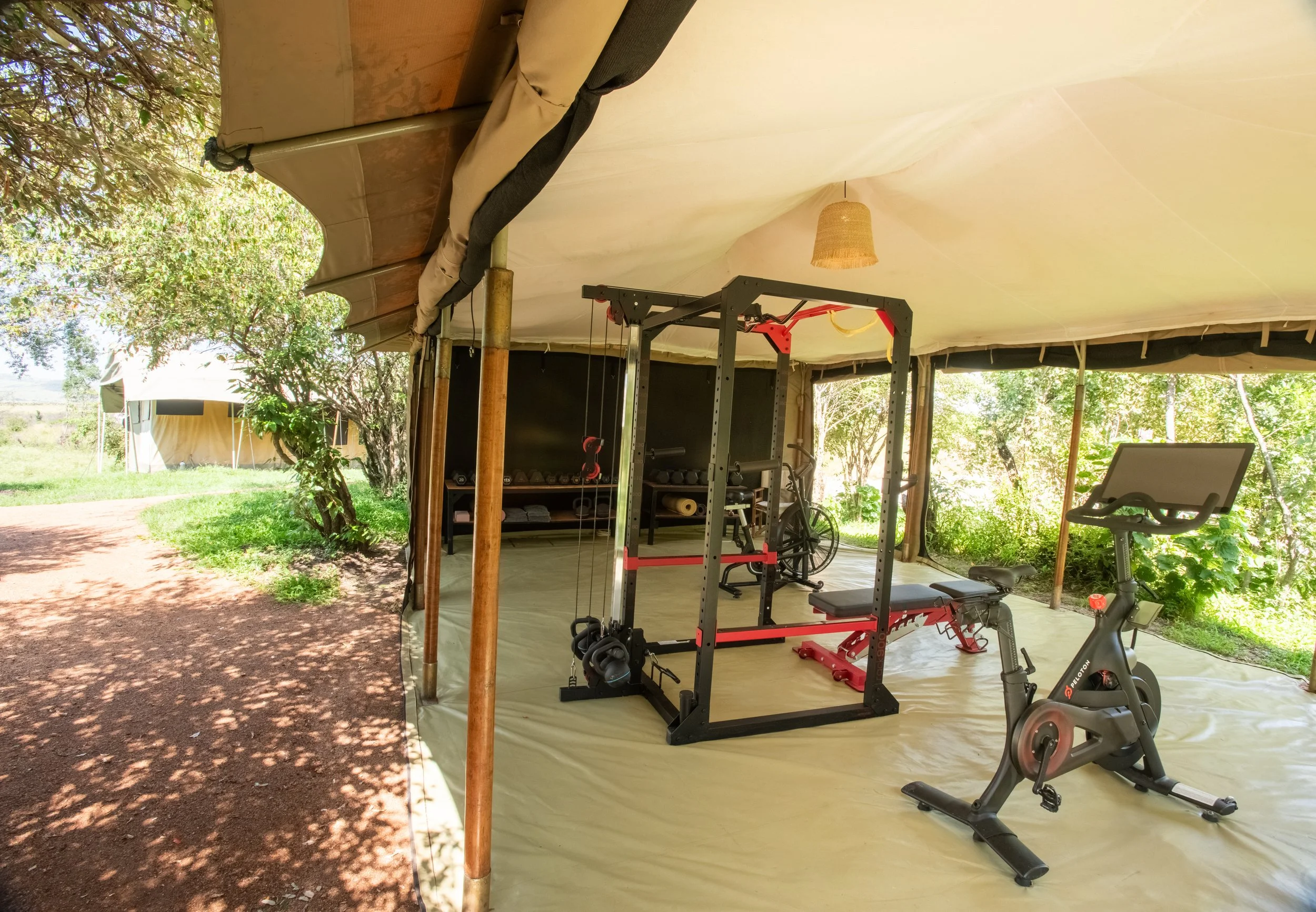 Exercise equipment inside a tent outdoors, including a stationary bike and a pull-up station, with trees and tents visible outside.