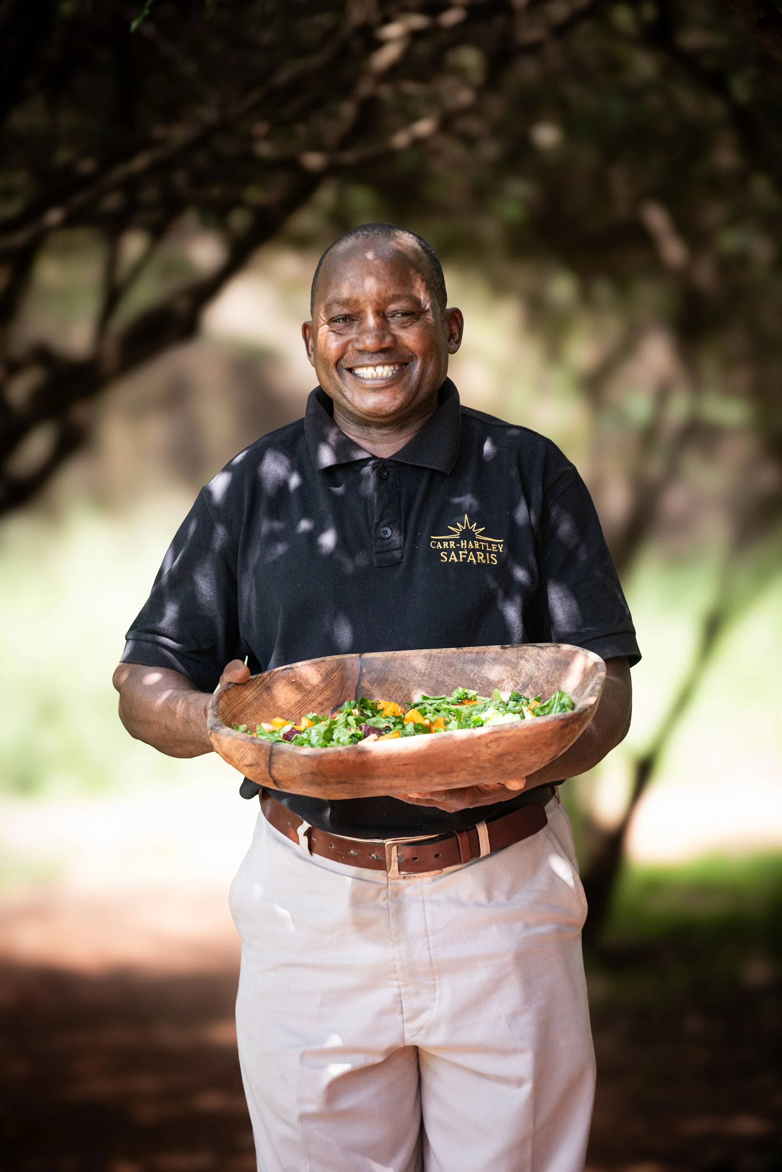 A smiling man in a black polo shirt with 'Carr-Hartley Safaris' logo, holding a wooden bowl filled with salad outdoors, with blurred trees in the background.