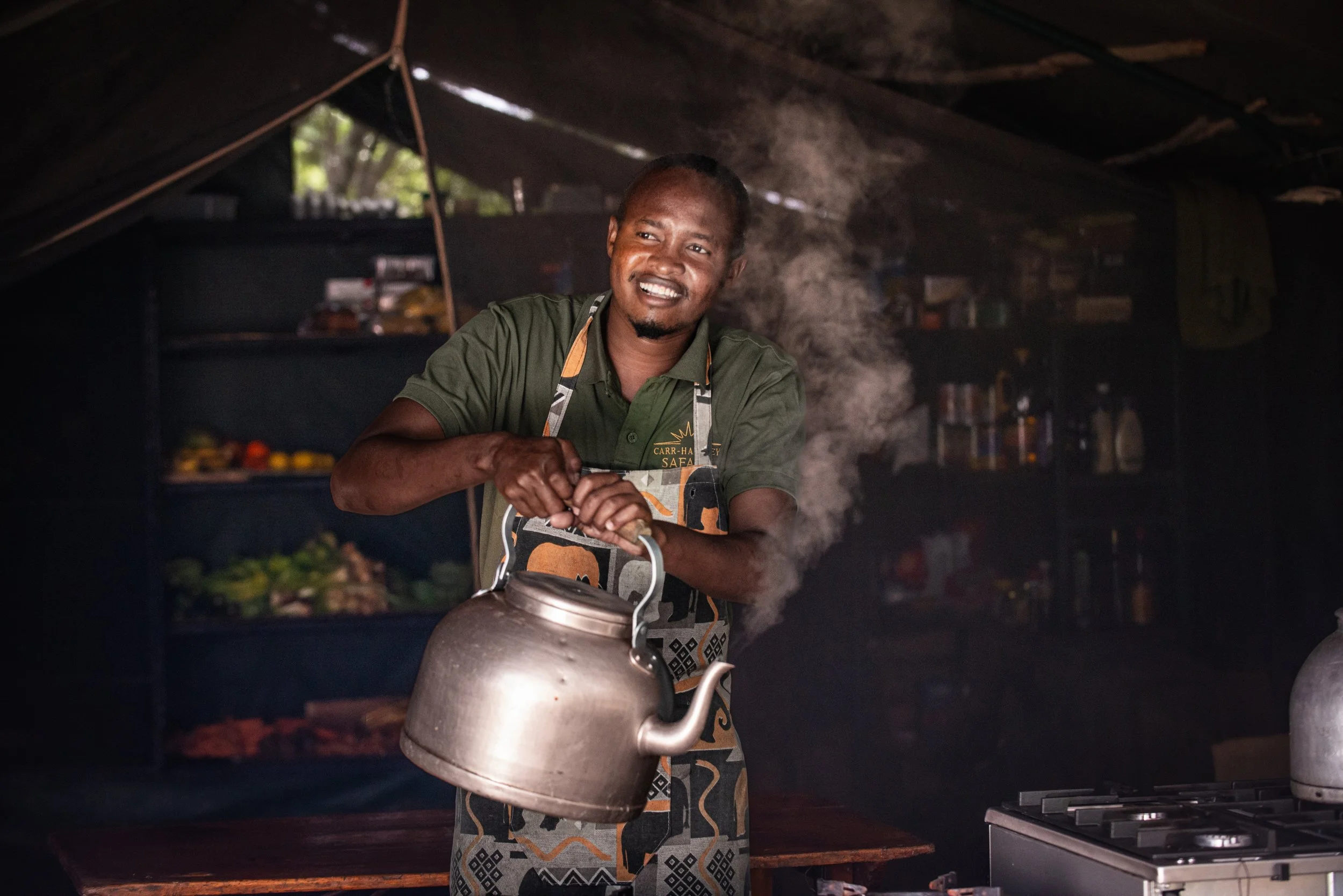 A man smiling and holding a kettle indoors, with steam rising from it, in a kitchen setting with shelves of food in the background.
