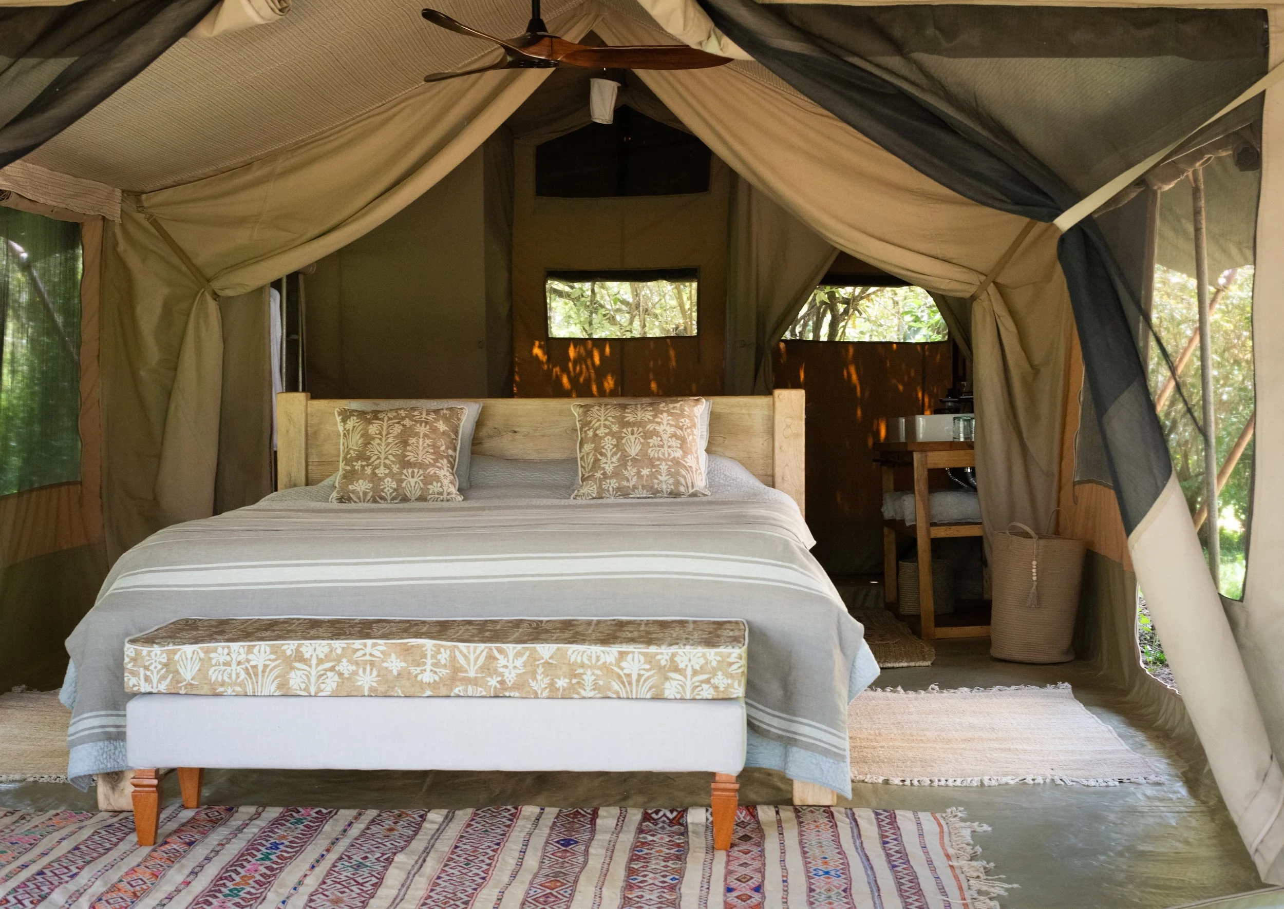Interior of a canvas tent with a large bed centered in the room, featuring beige and white striped bedding and two decorative pillows with floral patterns. At the foot of the bed is a small upholstered bench with a floral design. In the background, t