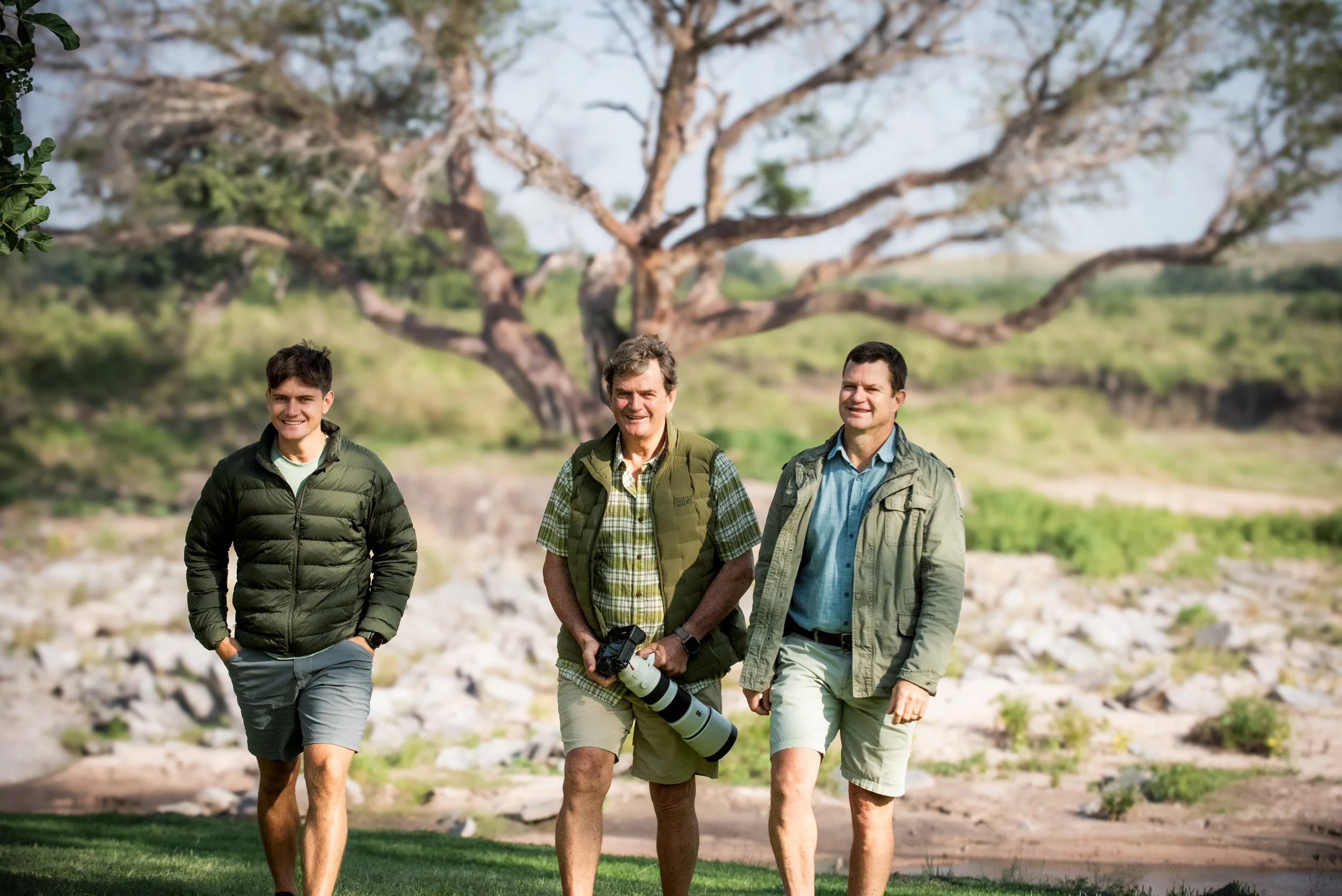 Three men walking outdoors in a natural setting with trees and rocky terrain, smiling and enjoying a sunny day.