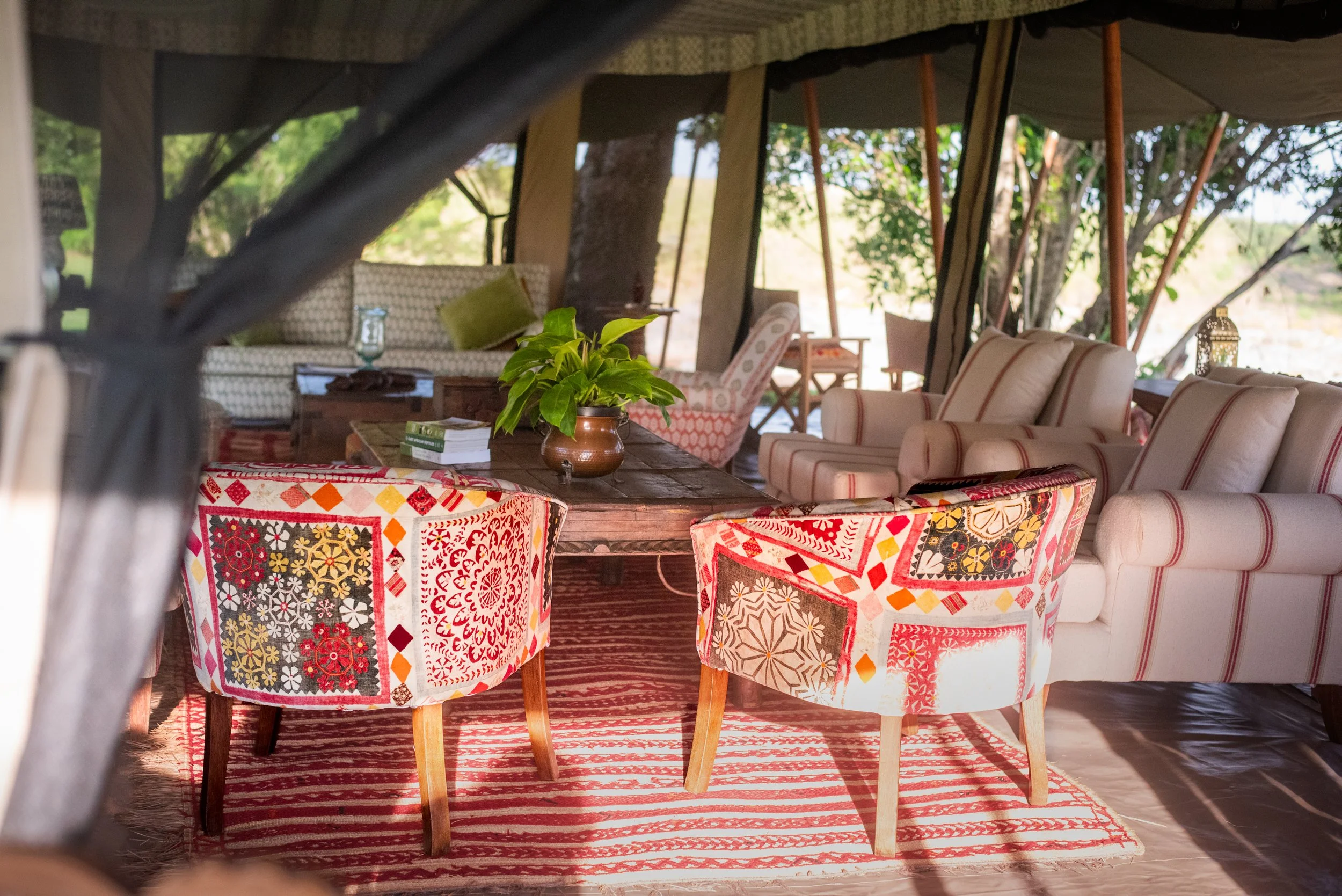 A cozy outdoor seating area with colorful patterned chairs around a wooden table, featuring a potted plant, on a striped rug, with couches and chairs in the background under a canopy in a natural setting.