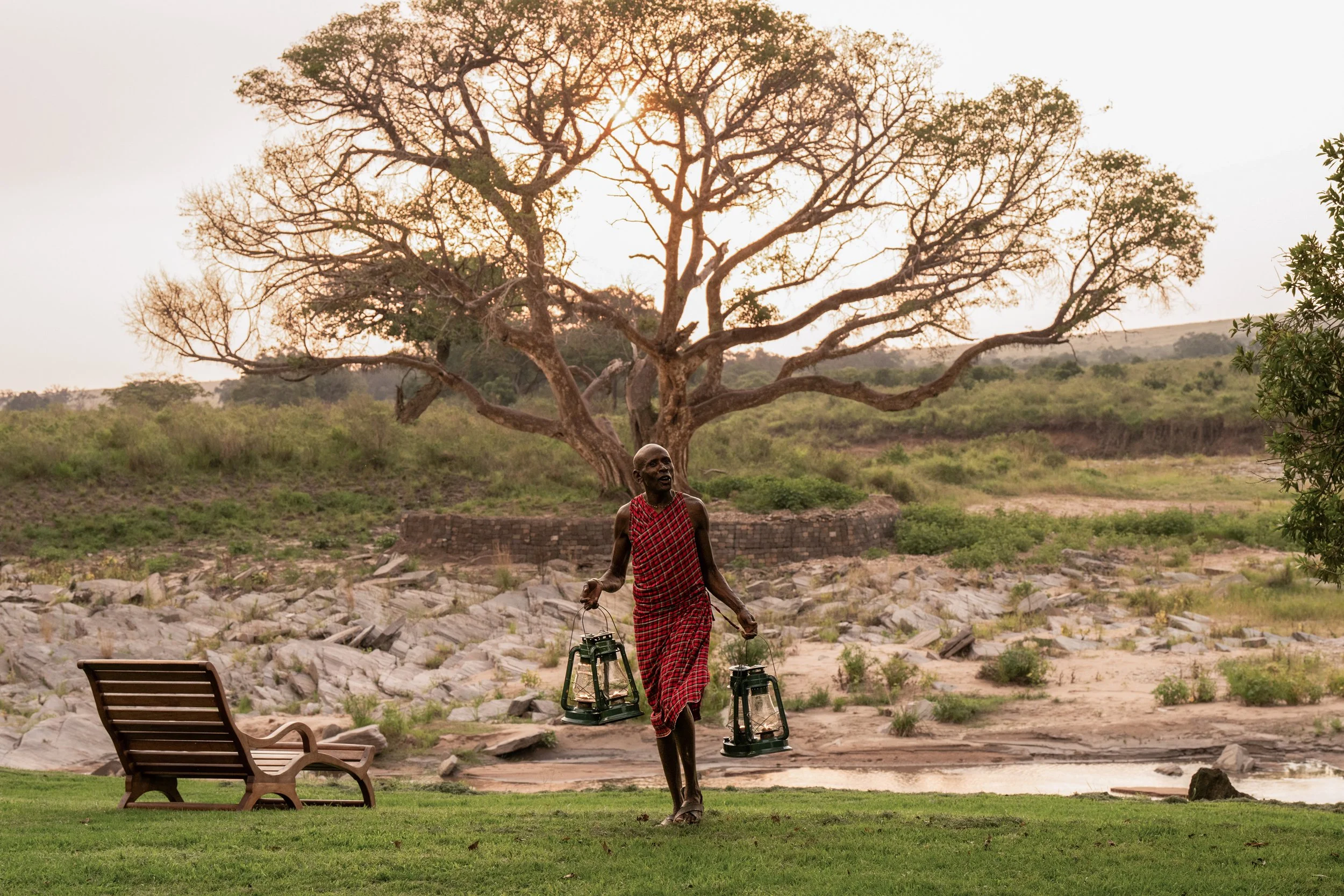 An elderly man with dark skin and a shaved head, dressed in traditional red and black patterned clothing, carrying two lanterns by their handles, walking across a grassy area near a river. In the background, there is a large leafless tree and green s