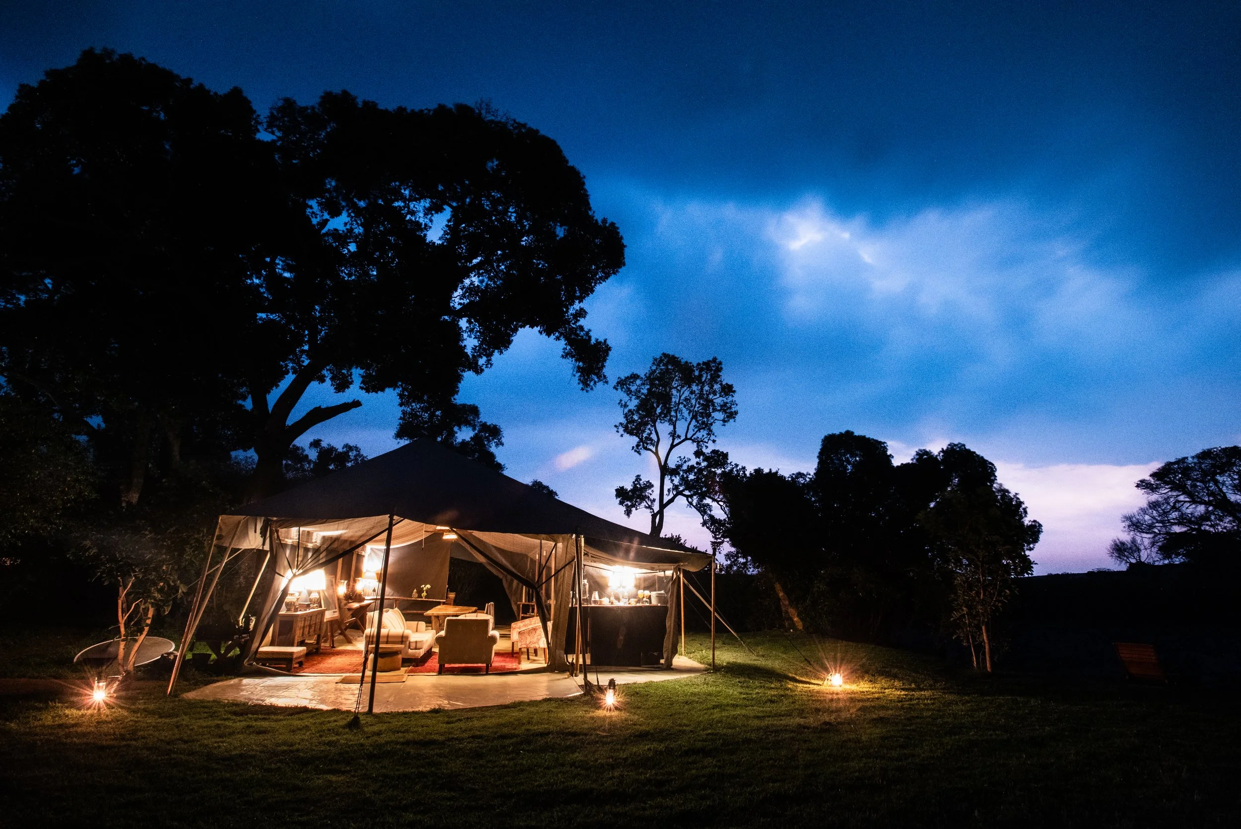 An outdoor tent set up in a grassy area during nighttime, illuminated with warm lights, with trees and a dark blue sky in the background.