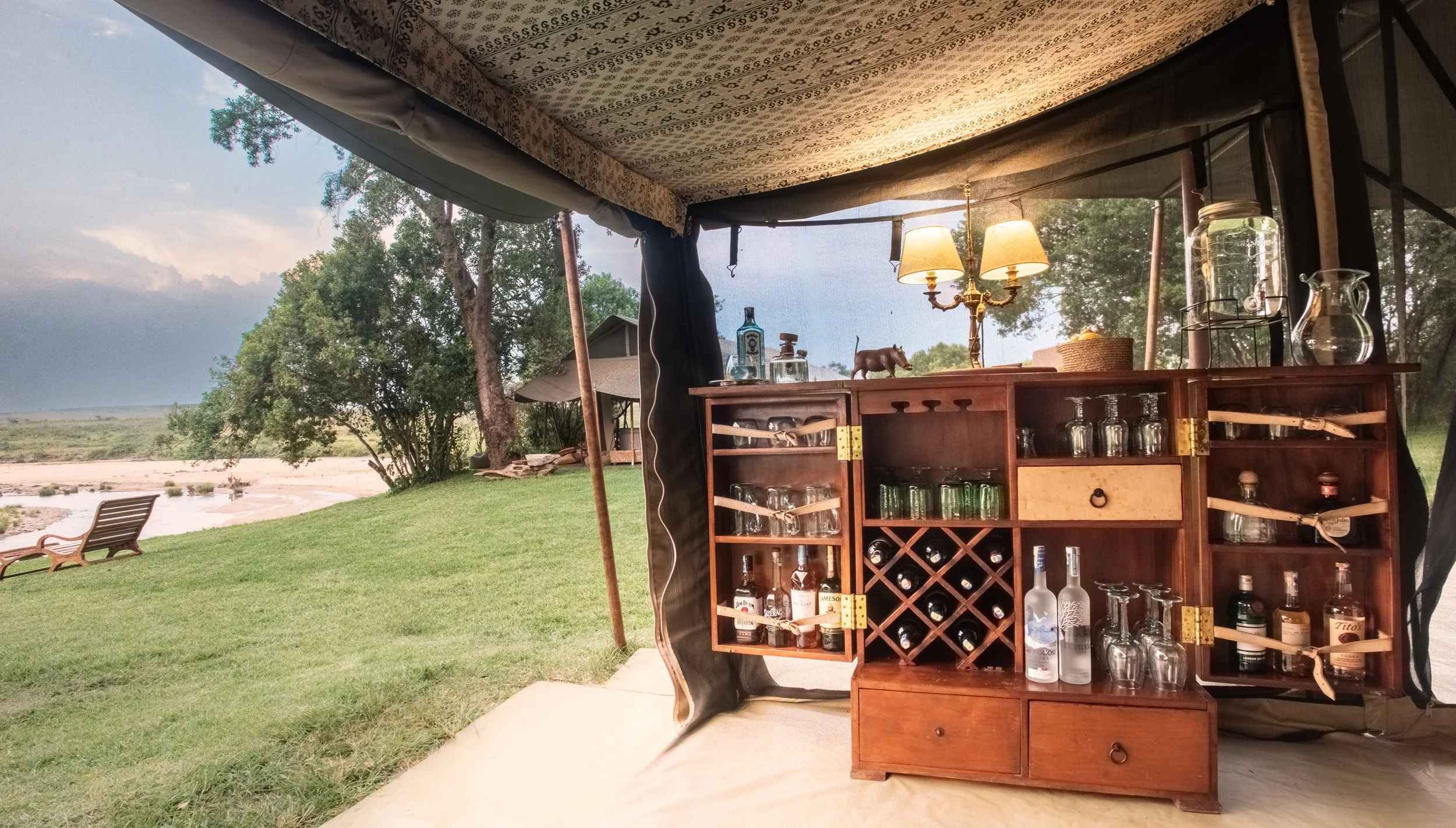 A cozy outdoor bar setup inside a tent or canopy with a wooden cabinet filled with liquor bottles and glassware. The background includes a grassy area with trees, a river, and lounge chairs facing the water, under a cloudy sky.