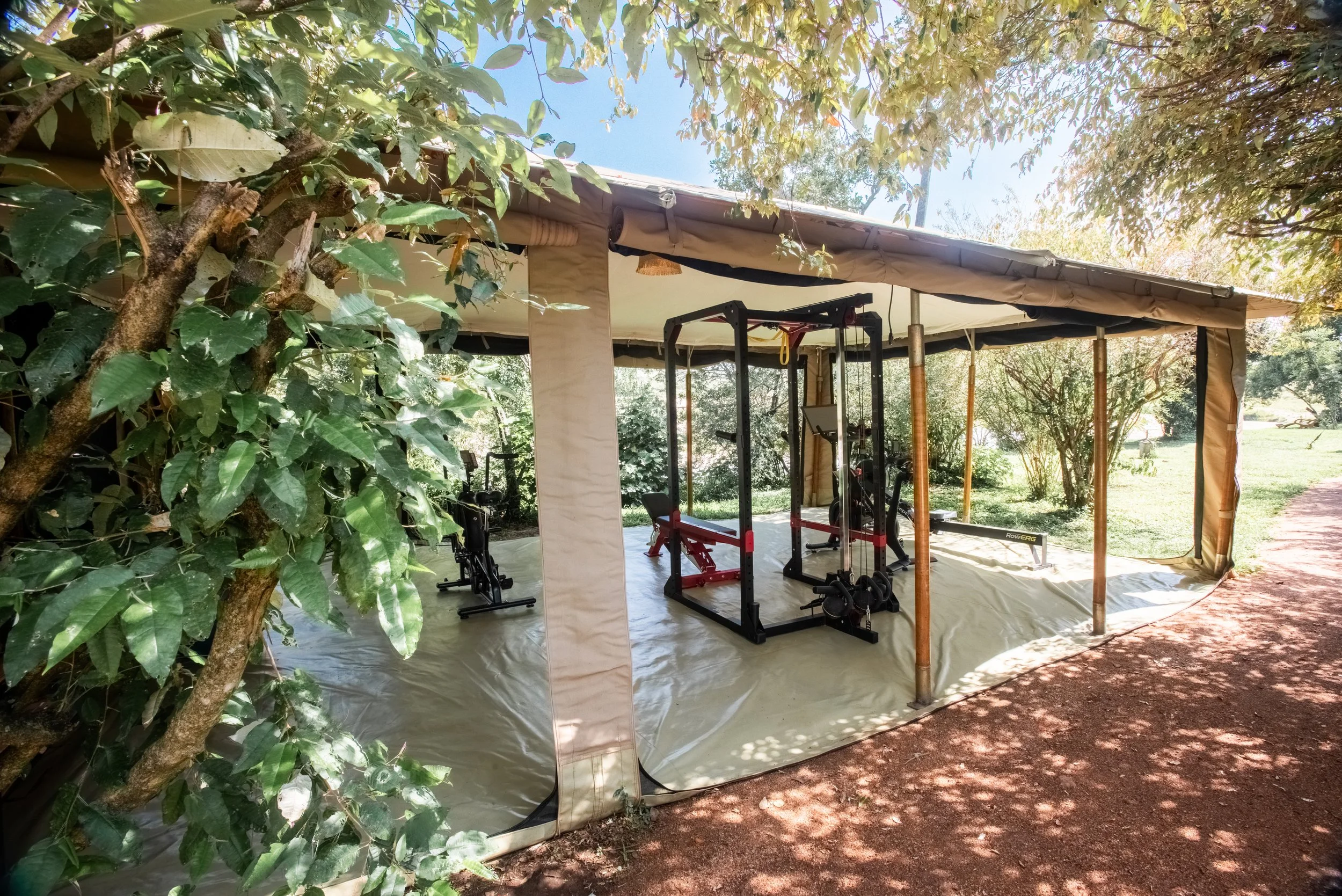A portable outdoor gym tent set up on the grass, containing gym equipment including a pull-up bar, a bench, and a workout bike, surrounded by trees and bushes with a dirt path nearby.