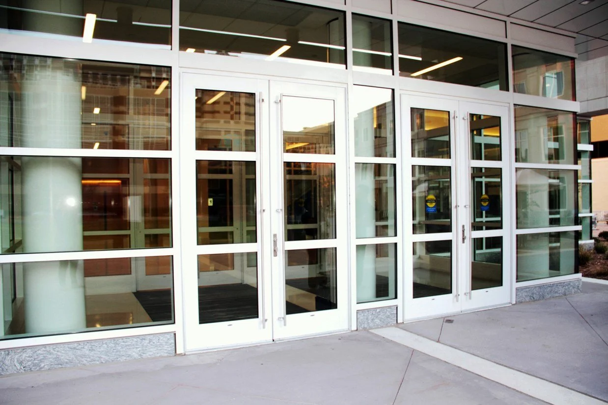 Front entrance of a modern building with glass doors and windows, metal framing, and concrete walkway.