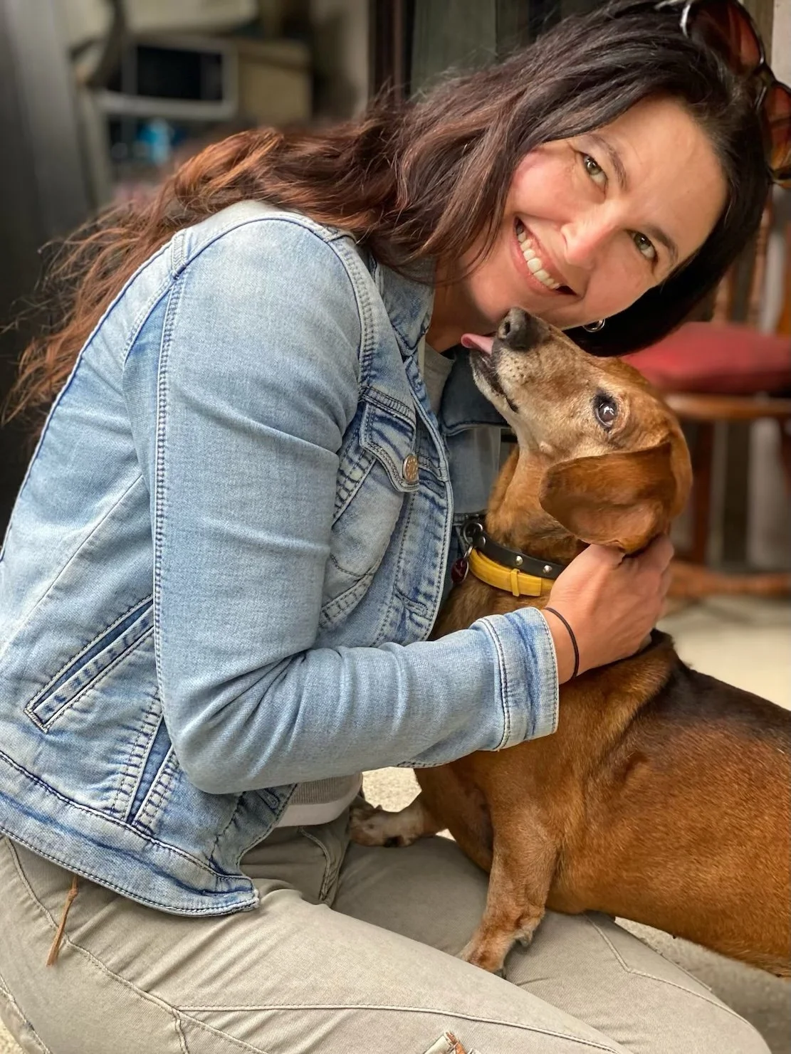 A woman with long brown hair, wearing a denim jacket, smiling while hugging a brown dachshund dog.