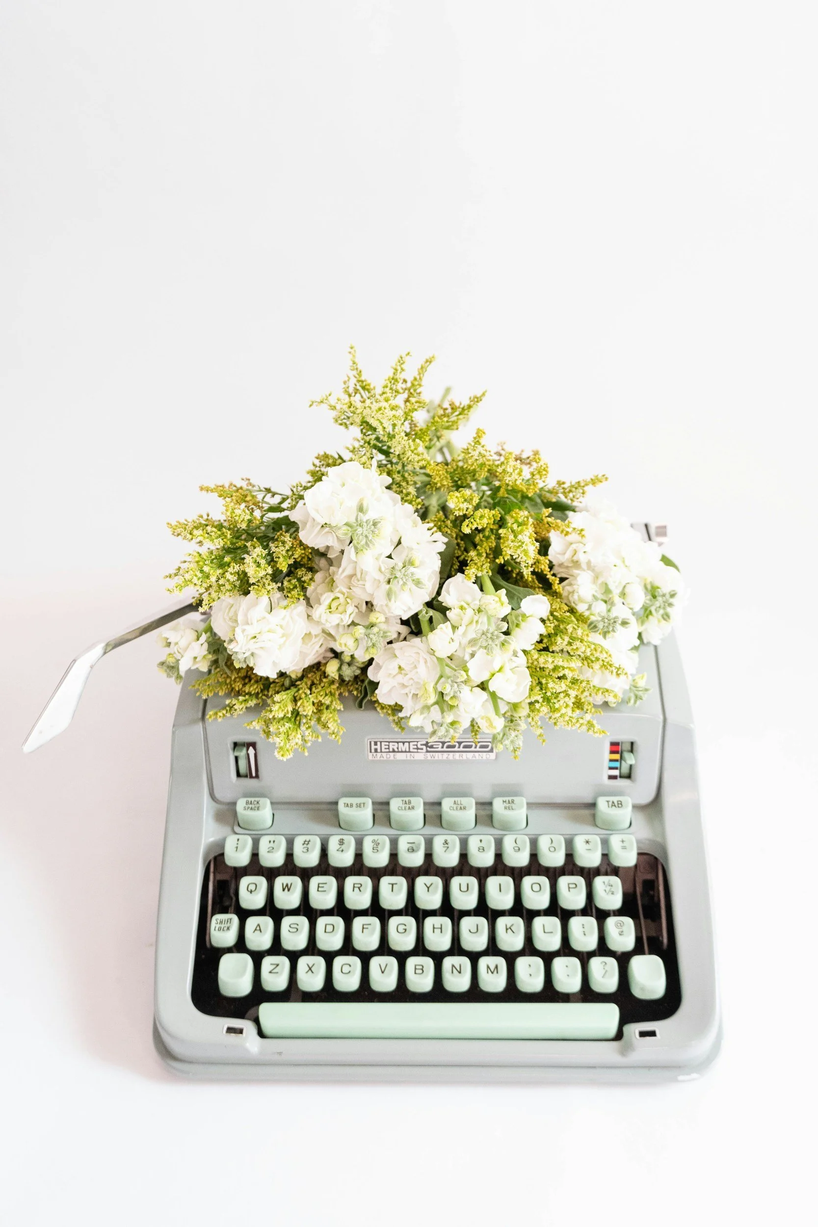 A vintage Hermes typewriter with white keys, adorned with a bouquet of white and yellow flowers.