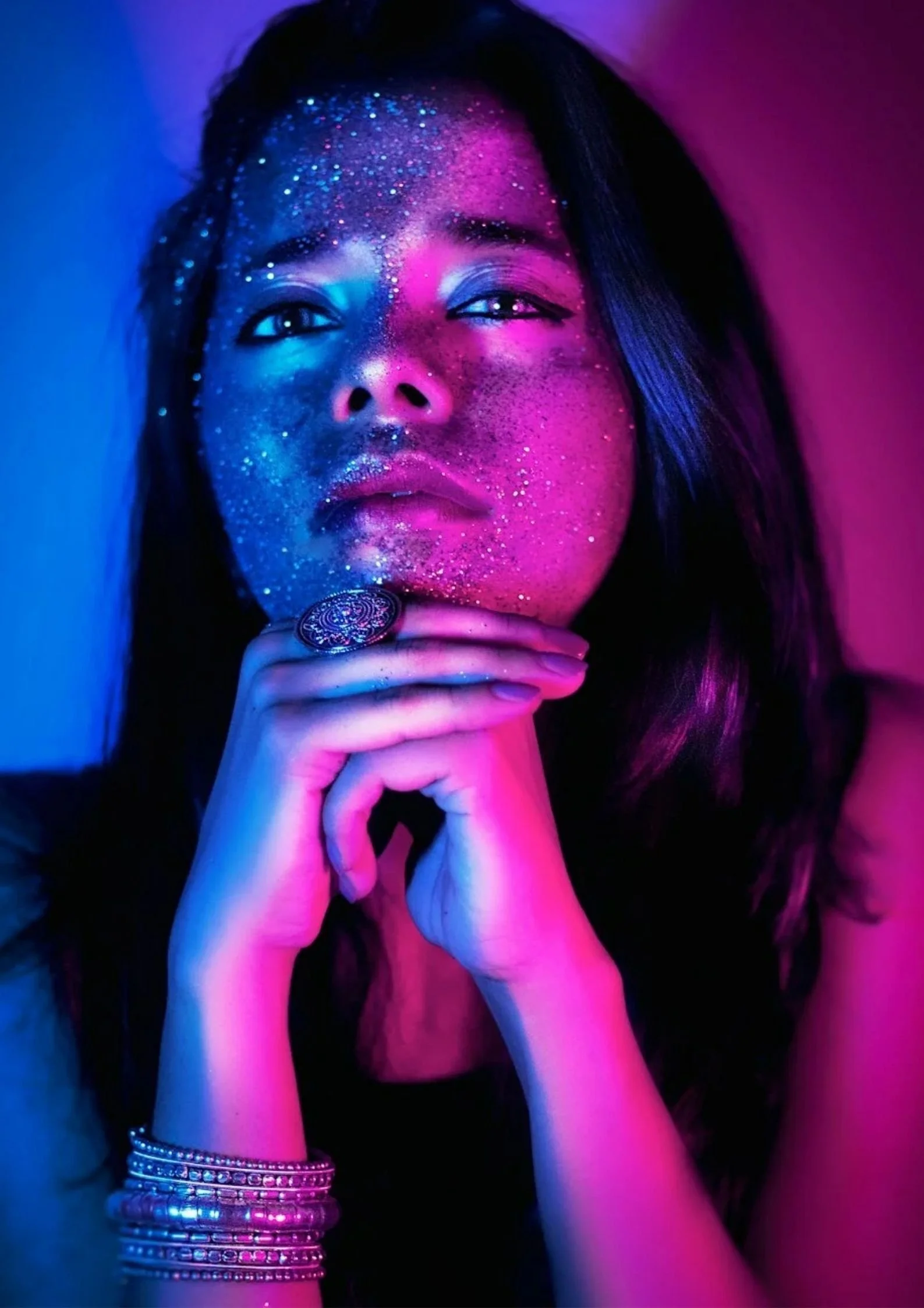 A woman with long dark hair and glitter makeup, illuminated by blue and pink neon lights, gazes at the camera with her hands clasped under her chin. She wears rings and stacked bracelets, and has glitter on her face and hands.