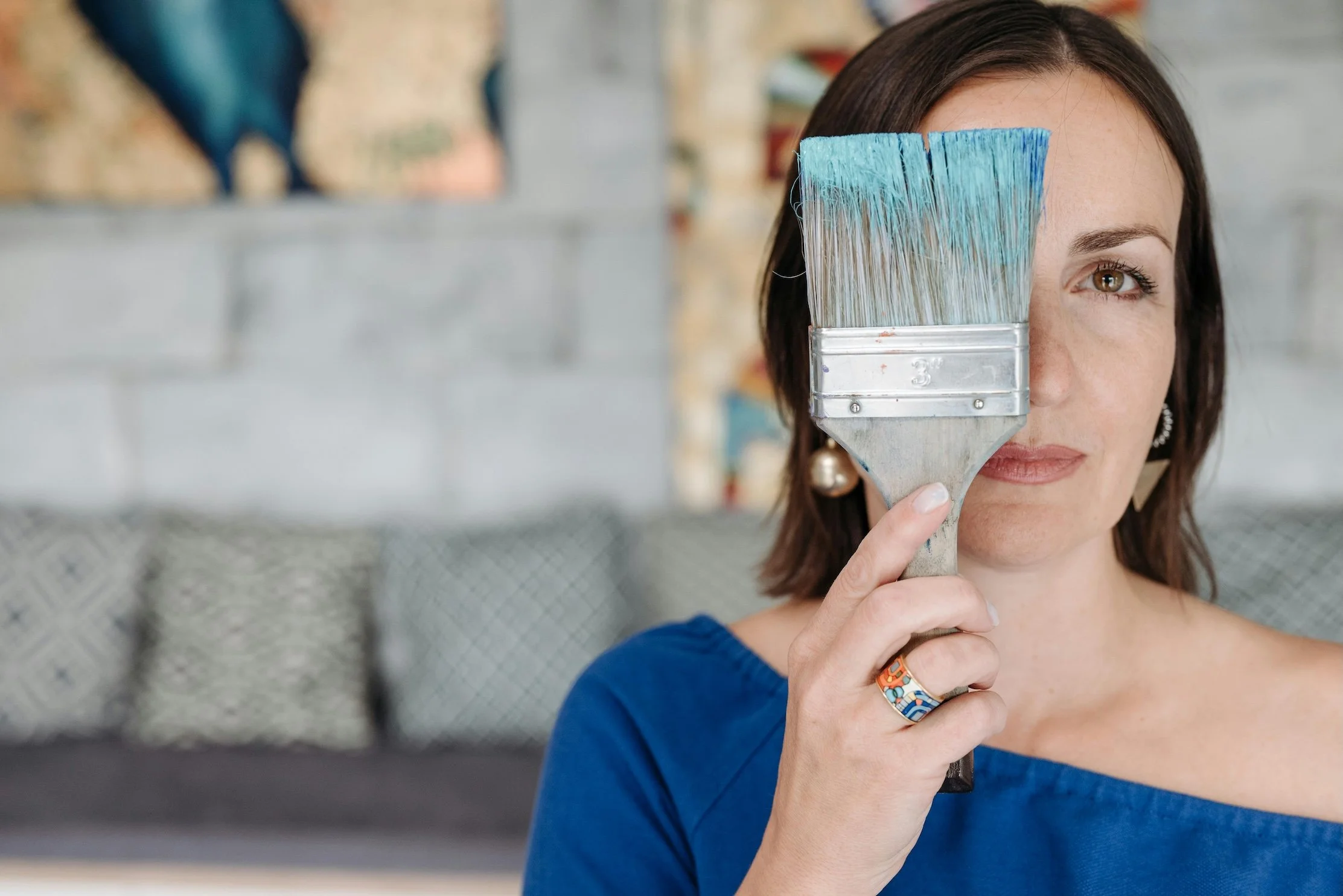 A woman with shoulder-length brown hair and earrings holding a paintbrush in front of her face, with the brush covering one eye, in an indoor setting with a brick wall background.