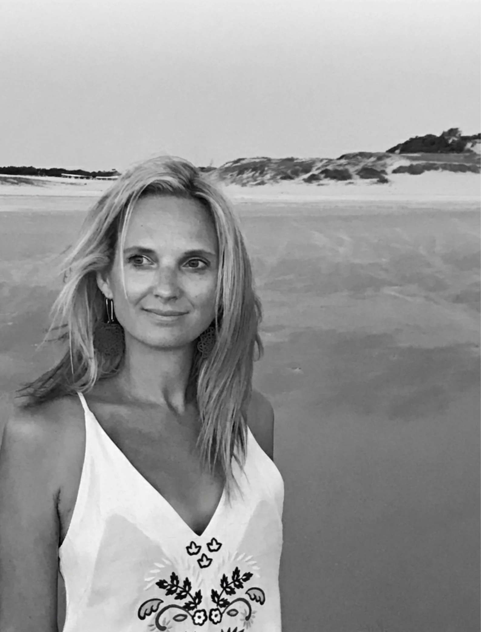 Portrait of website owner on beach in white dress with flower motif embroidery.