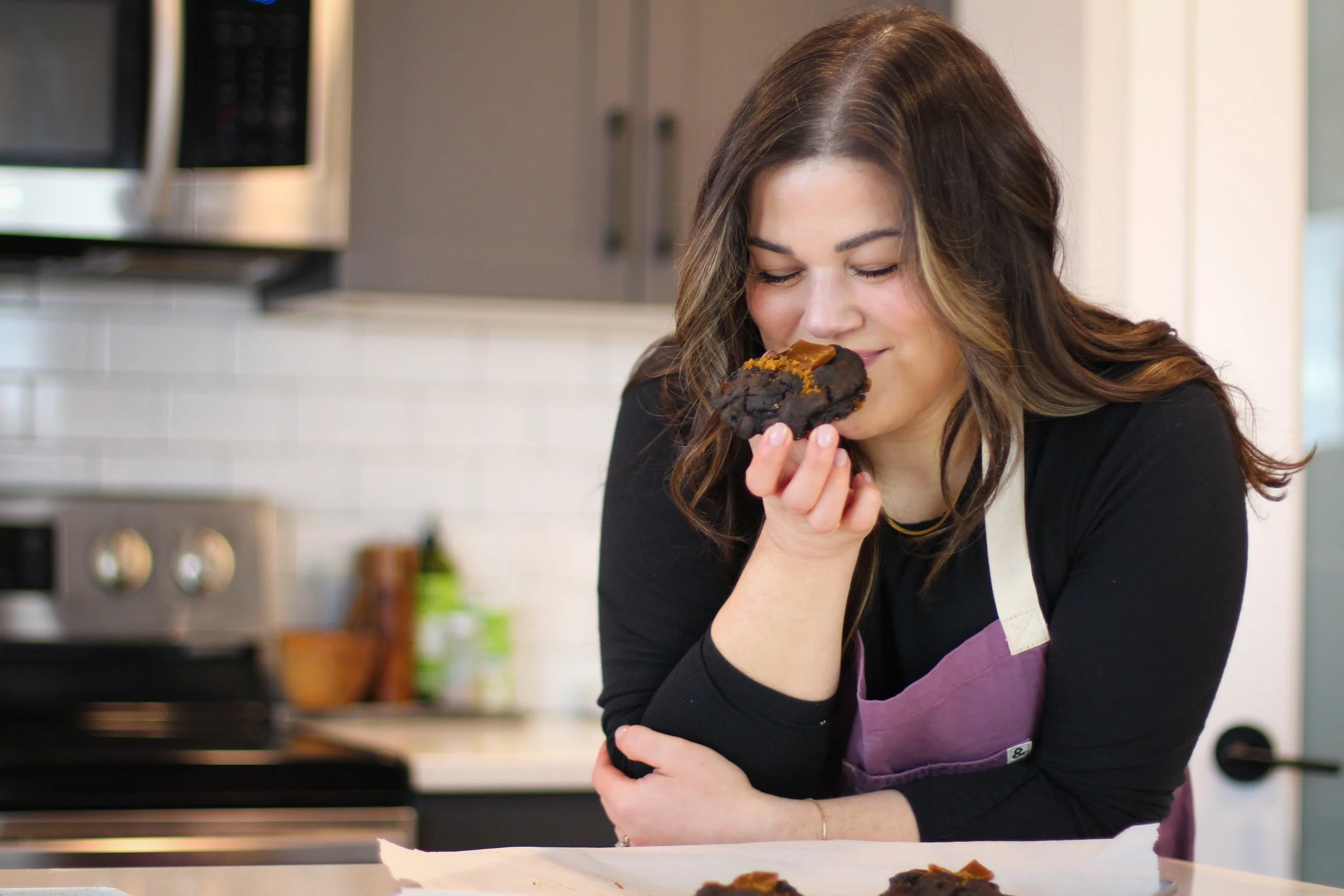A woman tasting a freshly baked chocolate cookie in a modern kitchen.