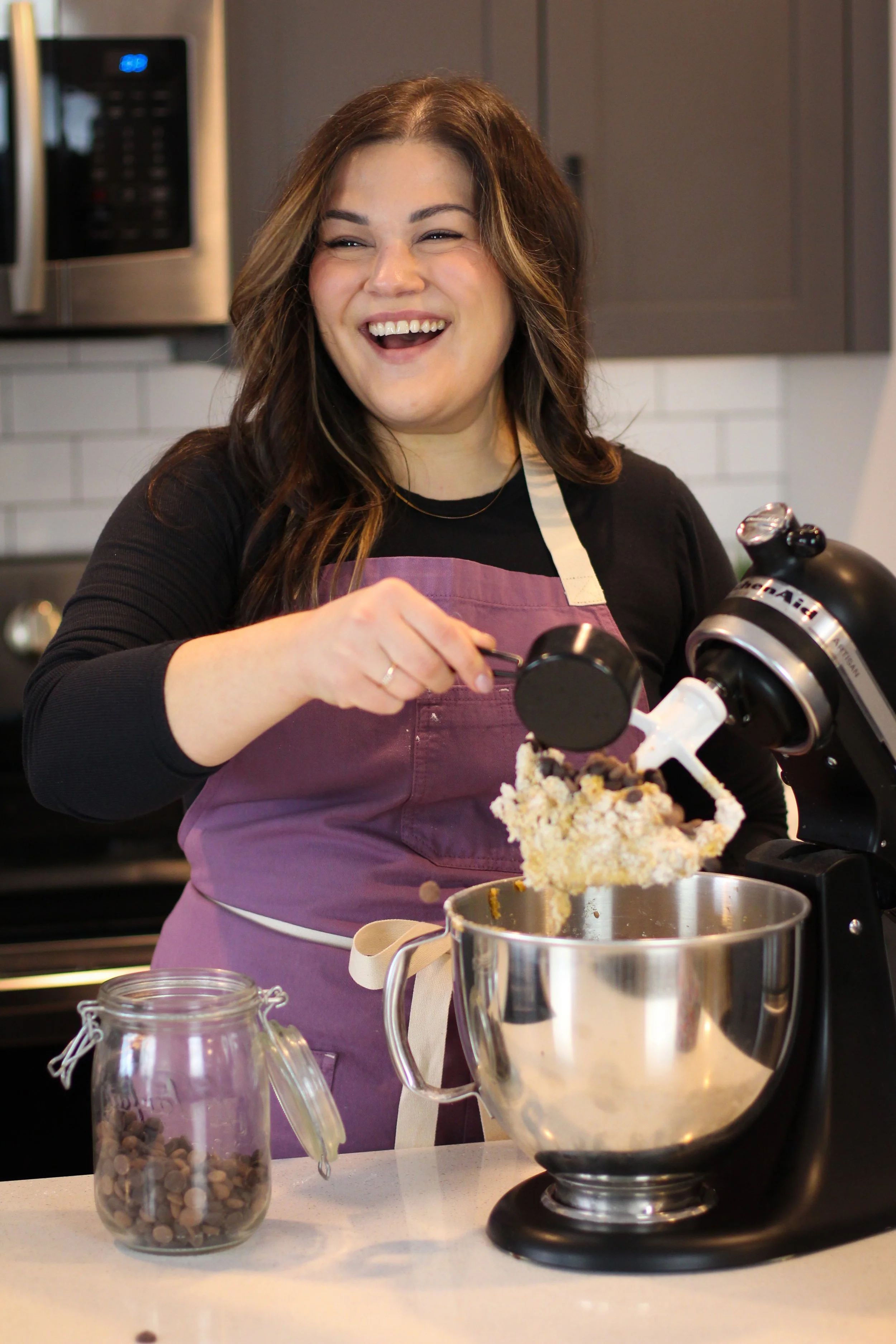 A woman wearing a pink apron smiles as she uses a stand mixer to prepare dough or batter in a kitchen.