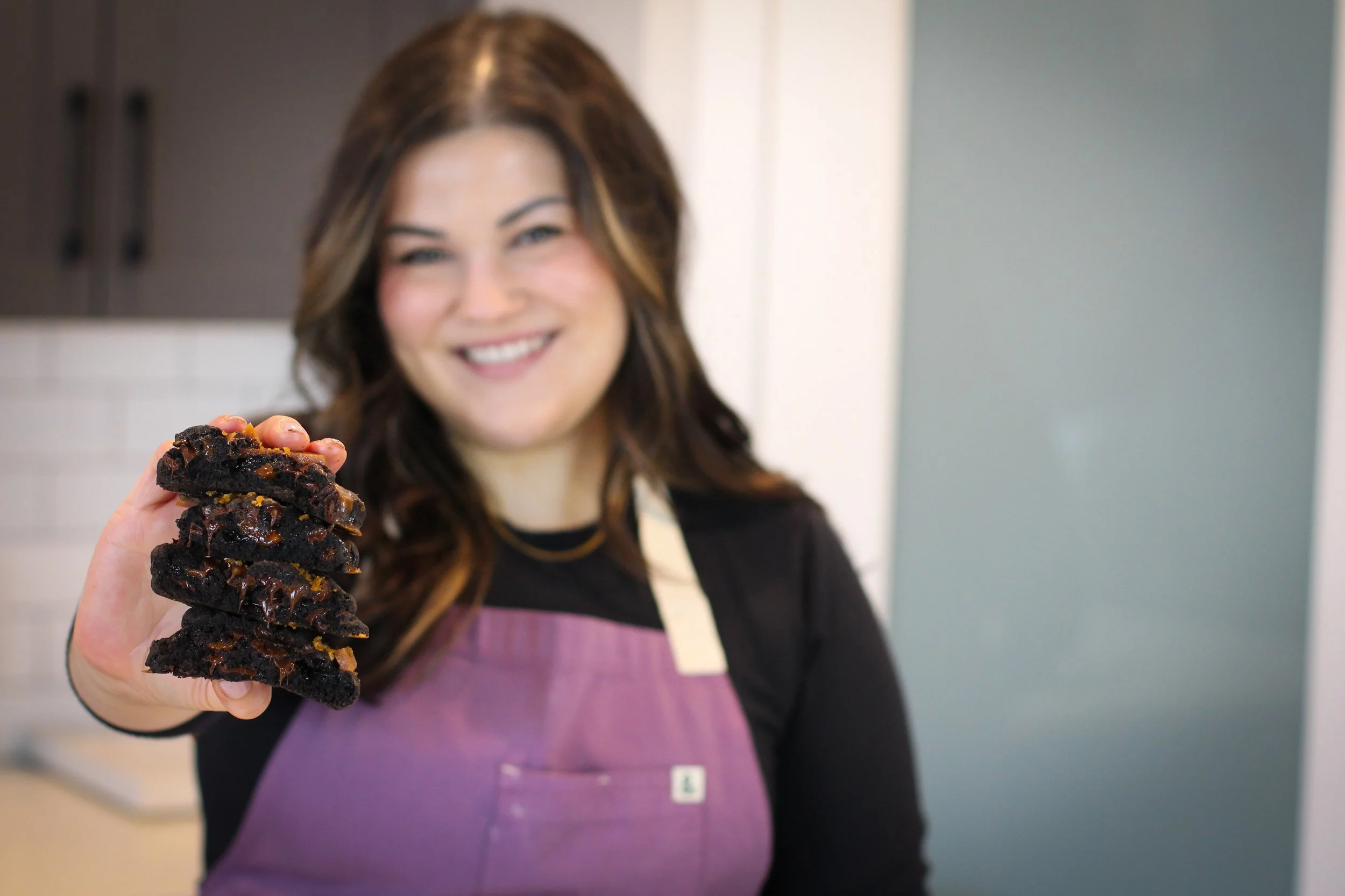 A woman with brown hair smiling and wearing a purple apron, holding a stack of chocolate cookies in a kitchen.