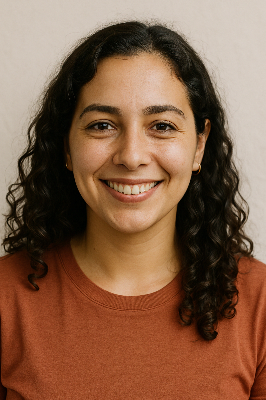 A smiling woman with curly dark hair, wearing a rust-colored shirt, standing against a neutral background.