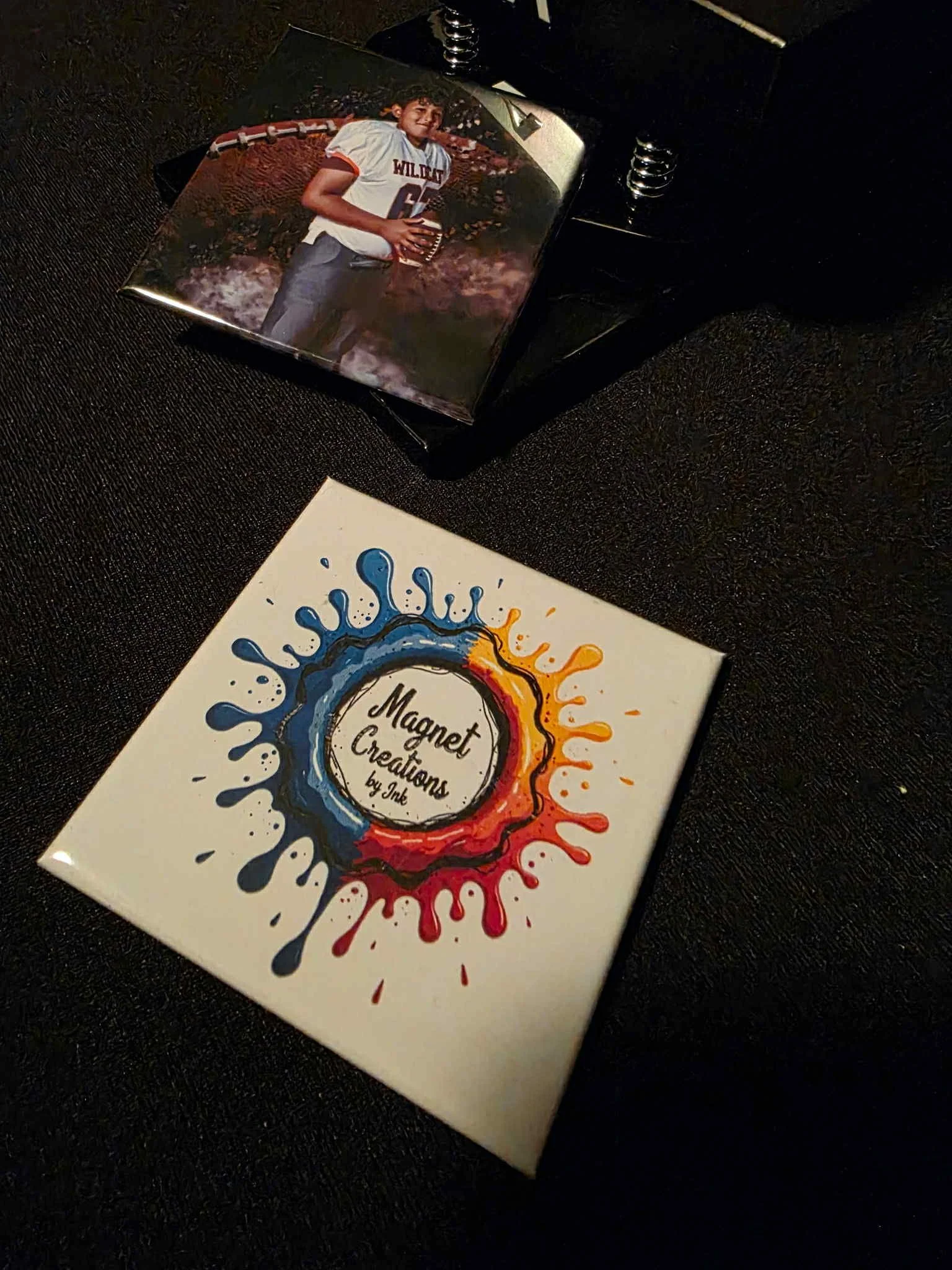 A square canvas with a colorful splatter design and the text 'Magnet Creations by Jne' in the center, placed on a black surface. Above it is a small photograph of a young boy in a white jersey holding a football.