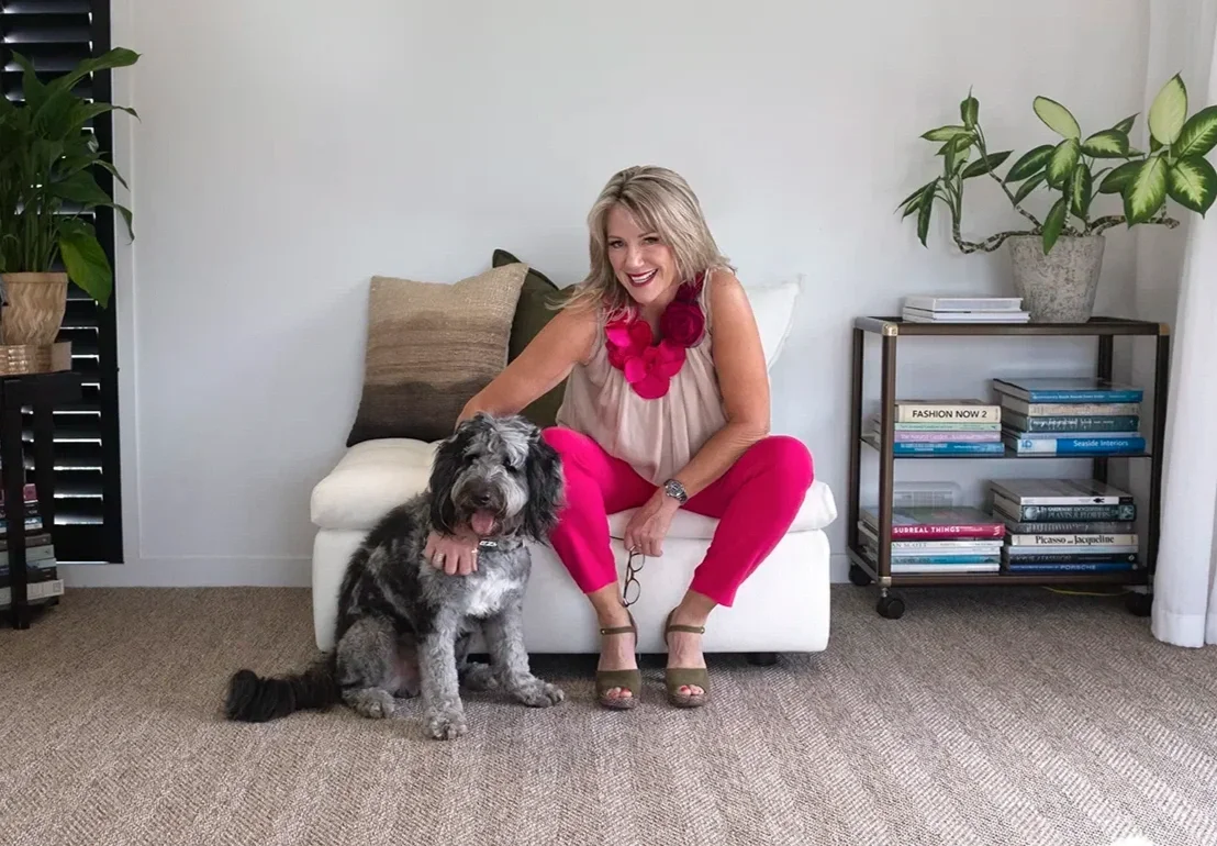 Woman in pink pants and beige top sitting on a white sofa, smiling, with a large, fluffy black and gray dog sitting beside her, in a living room with bookshelves and potted plants.