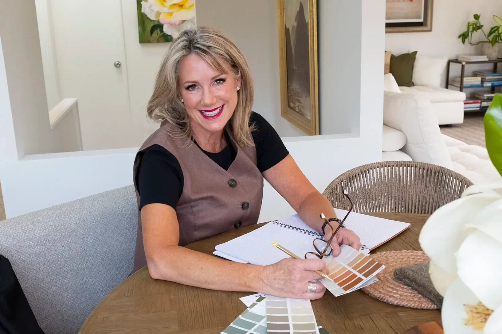 A smiling woman with blonde hair, wearing a black shirt and a brown vest, sitting at a wooden table with color swatches, a notebook, and glasses in her hands, in a cozy, well-decorated living space.