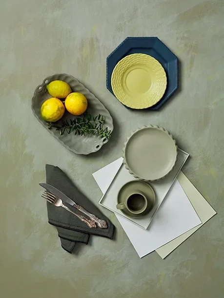 Overhead view of a table setting with yellow lemons and greenery on an oval gray dish, stacked dinner plates in yellow and blue, a gray bowl on white paper, a gray mug, silverware on a dark gray napkin, and blank white and beige papers on a light green surface.