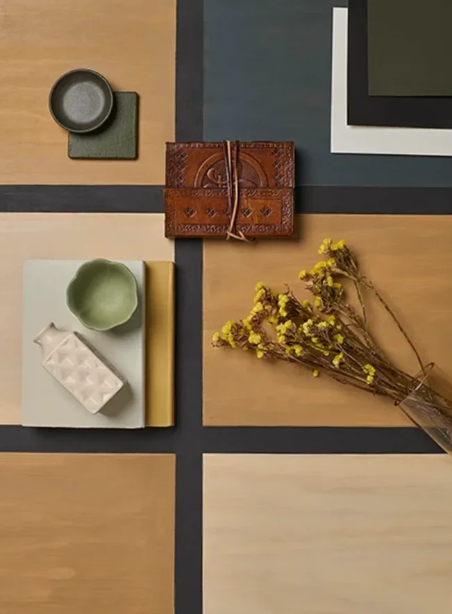 Flat lay of various objects on a wooden and tiled surface, including a small dark dish, a square patch of fabric, a wallet, a small green bowl, a patterned white soap, a slim yellow notebook, and a bunch of yellow flowers in a glass container.