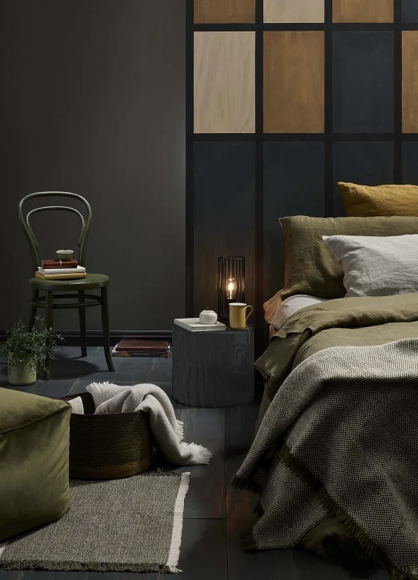 A cozy bedroom featuring a bed with beige and white bedding, a bedside table with a small lamp, a green chair with books, a potted plant, and a dark accent wall with geometric panels.