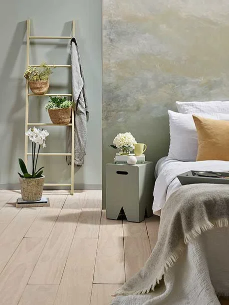 A bedroom corner with a bed, a light green nightstand with white flowers, a potted orchid, and a decorative ladder with hanging plants and a towel.