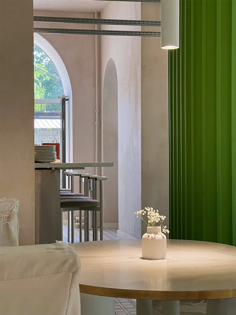 Interior of a modern room featuring a round wooden table with a white vase of small white flowers, bar stools, a large window with an arched top, and green vertical slats on a wall.