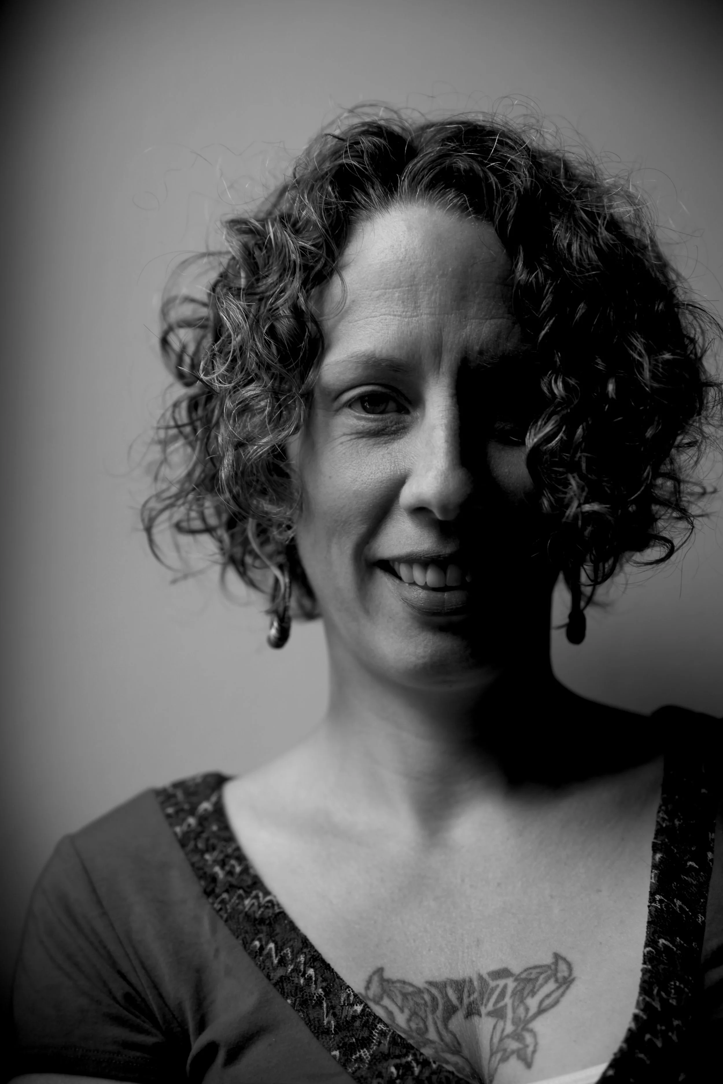 Black and white portrait of a smiling woman with curly hair, wearing a top with a decorative neckline, earrings, and a butterfly tattoo on her chest.