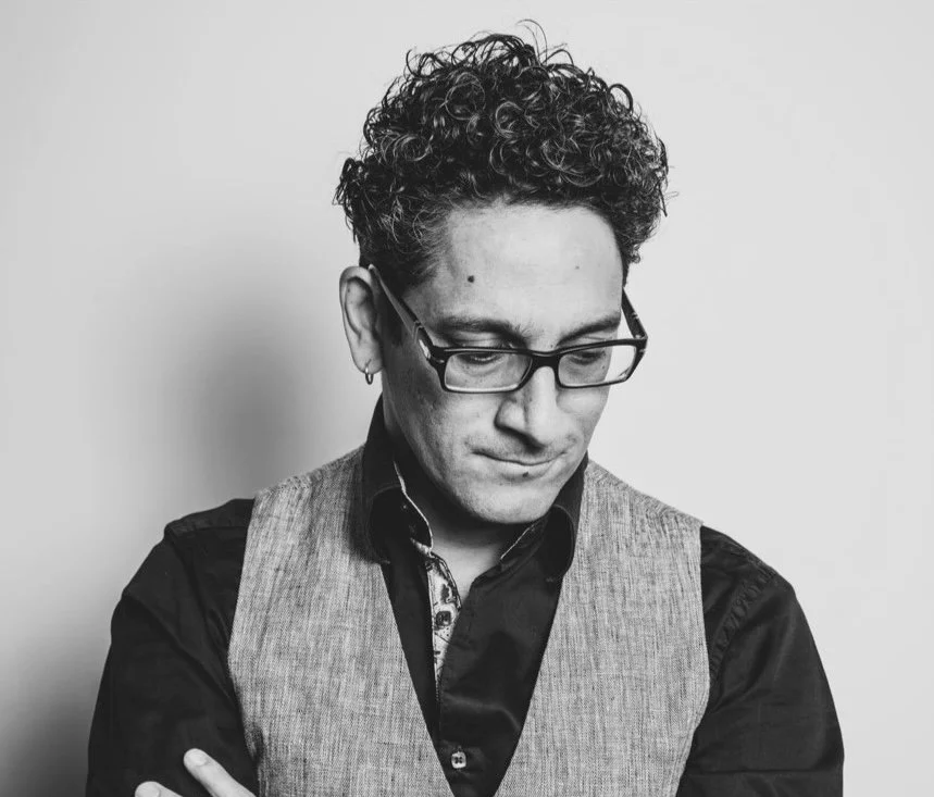 Black and white portrait of a man with glasses, curly hair, wearing a vest over a shirt, looking downward.