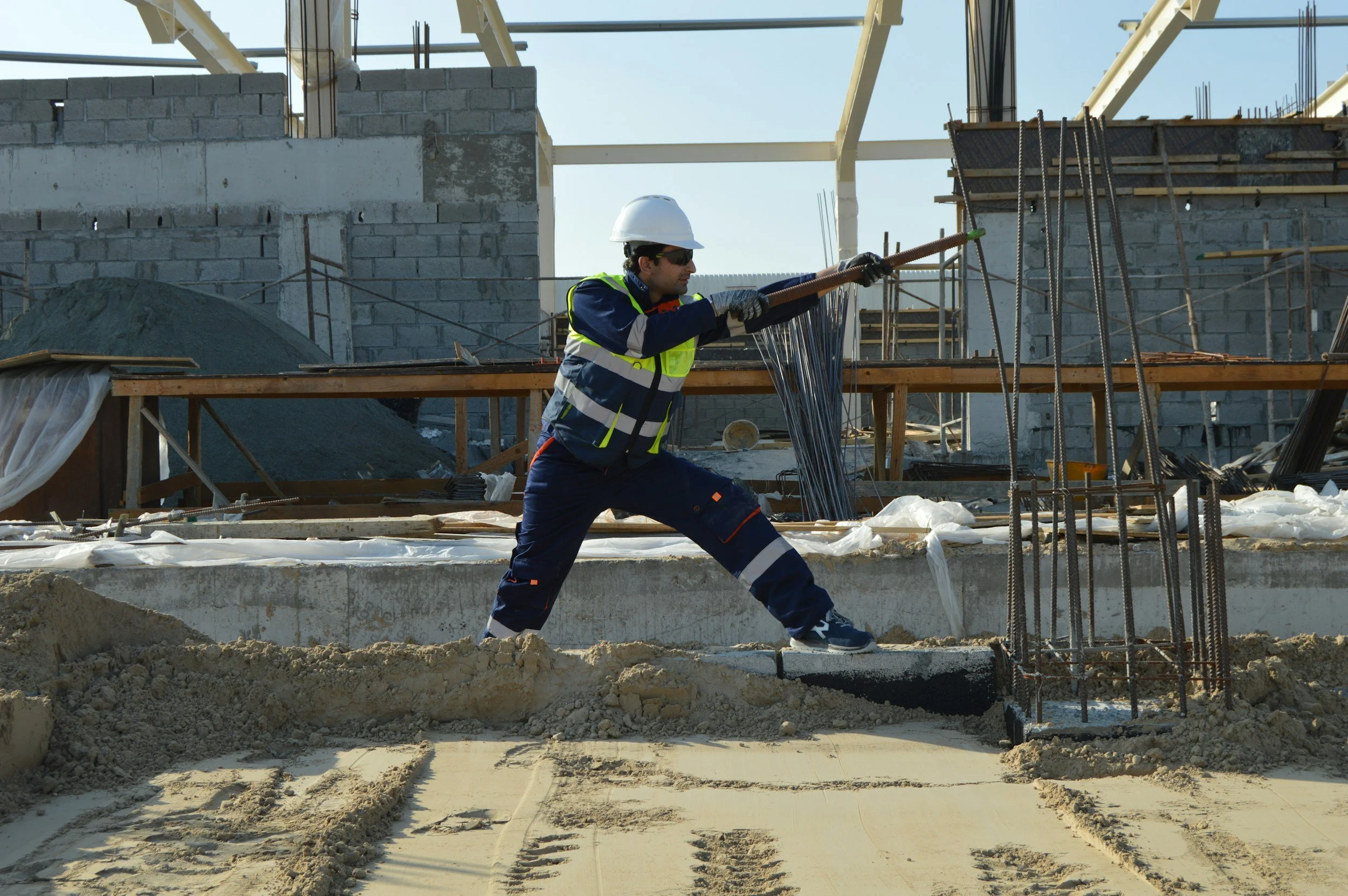 Construction worker in safety gear, including a helmet and reflective vest, using a tool on rebar at a building site with unfinished concrete walls and scaffolding.